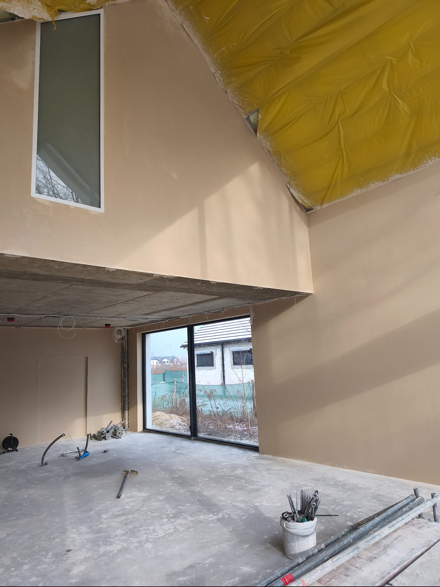 Wnętrze po tynkowaniu maszynowym: gładkie, beżowe ściany, duże okna z widokiem na zewnątrz, żółta folia zabezpieczająca sufit, narzędzia na podłodze.