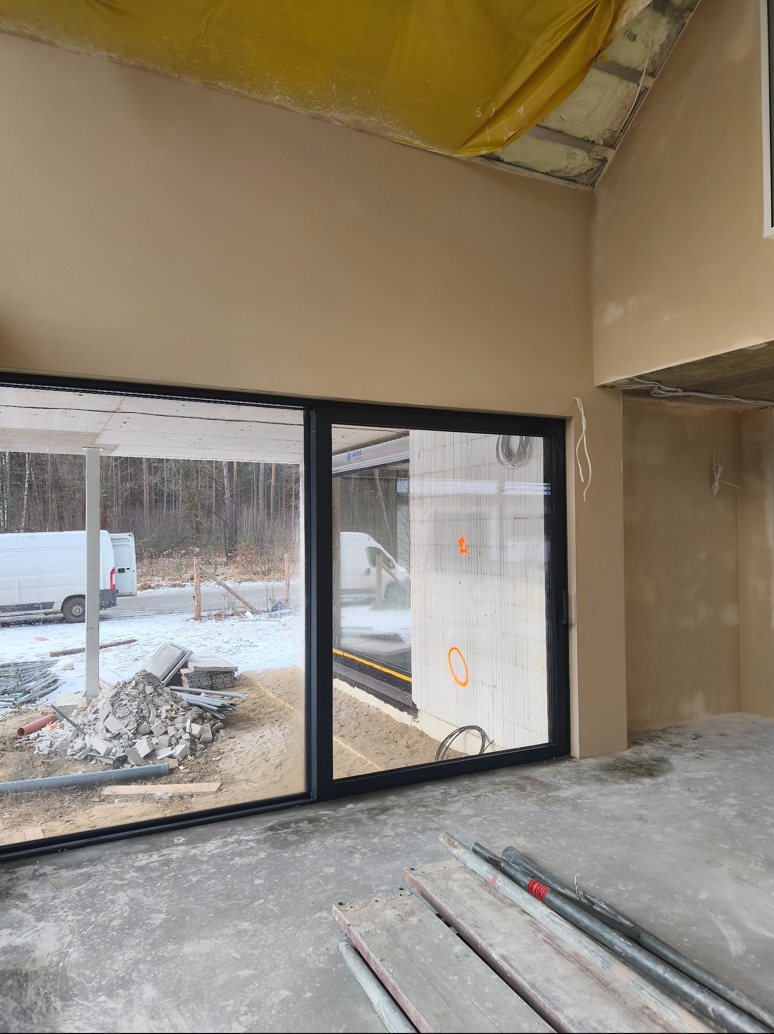 Wnętrze w trakcie prac wykończeniowych: ściany po nałożeniu tynku maszynowego, widoczne okno z widokiem na zewnątrz i elementy rusztowania w pomieszczeniu.