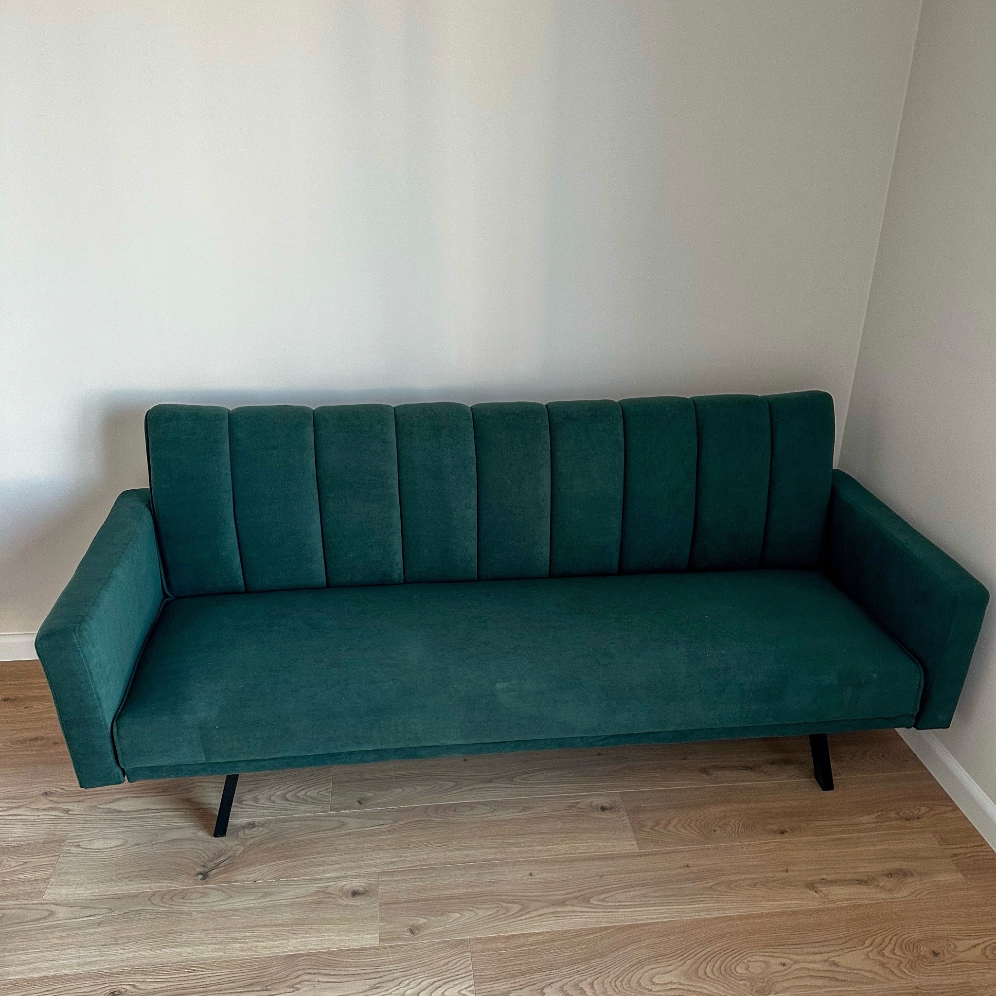 Szmaragdowa sofa z weluru na drewnianej podłodze, ustawiona pod białą ścianą. Minimalistyczny design z pionowymi przeszyciami na oparciu i czarnymi nogami.