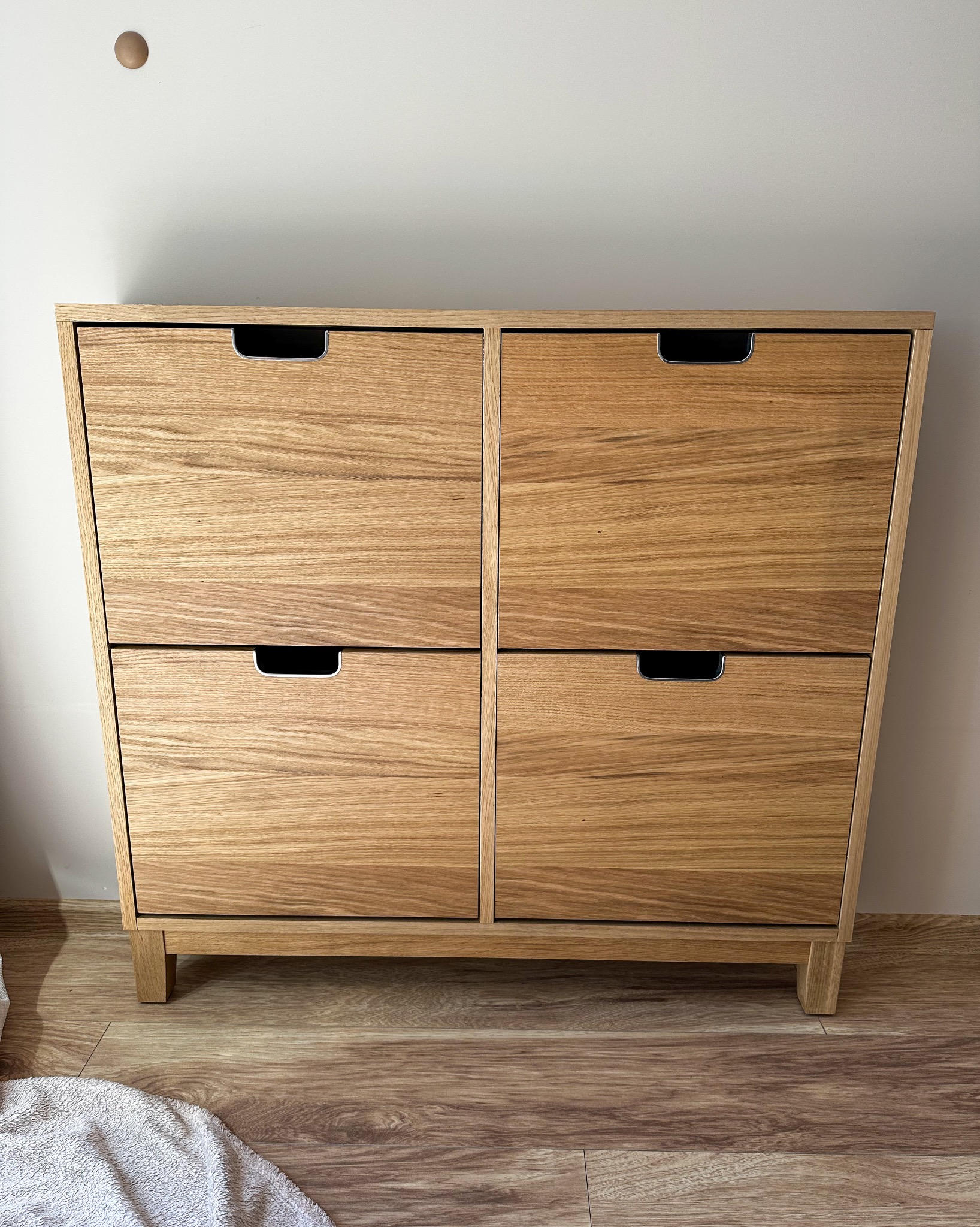 Czterodrzwiowa komoda z jasnego drewna z metalowymi uchwytami, ustawiona na drewnianej podłodze, z widocznym fragmentem białego ręcznika.