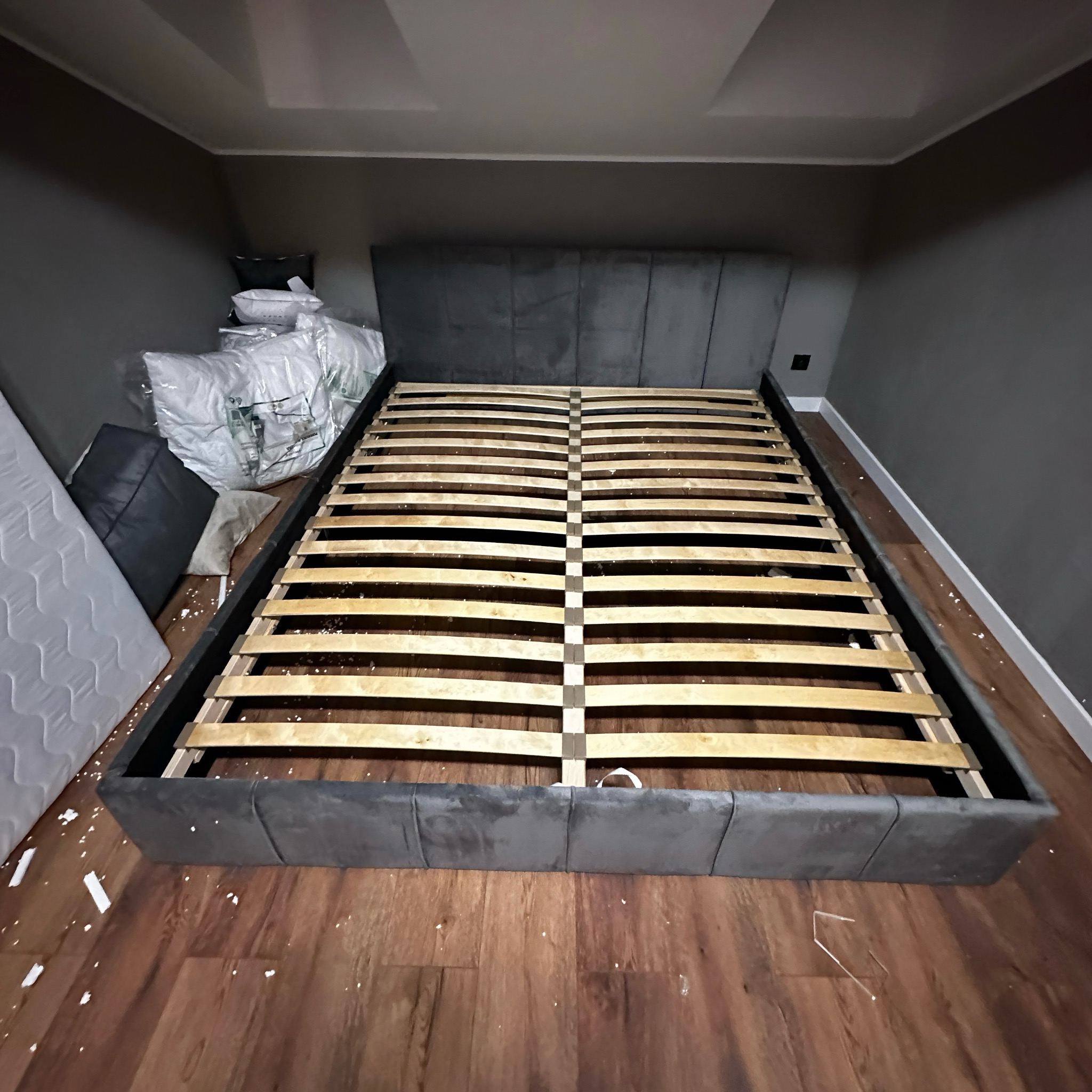 Szare łóżko z drewnianym stelażem w trakcie montażu. Widoczne elementy montażowe na podłodze. W tle ściana w odcieniach szarości i białe poduszki.