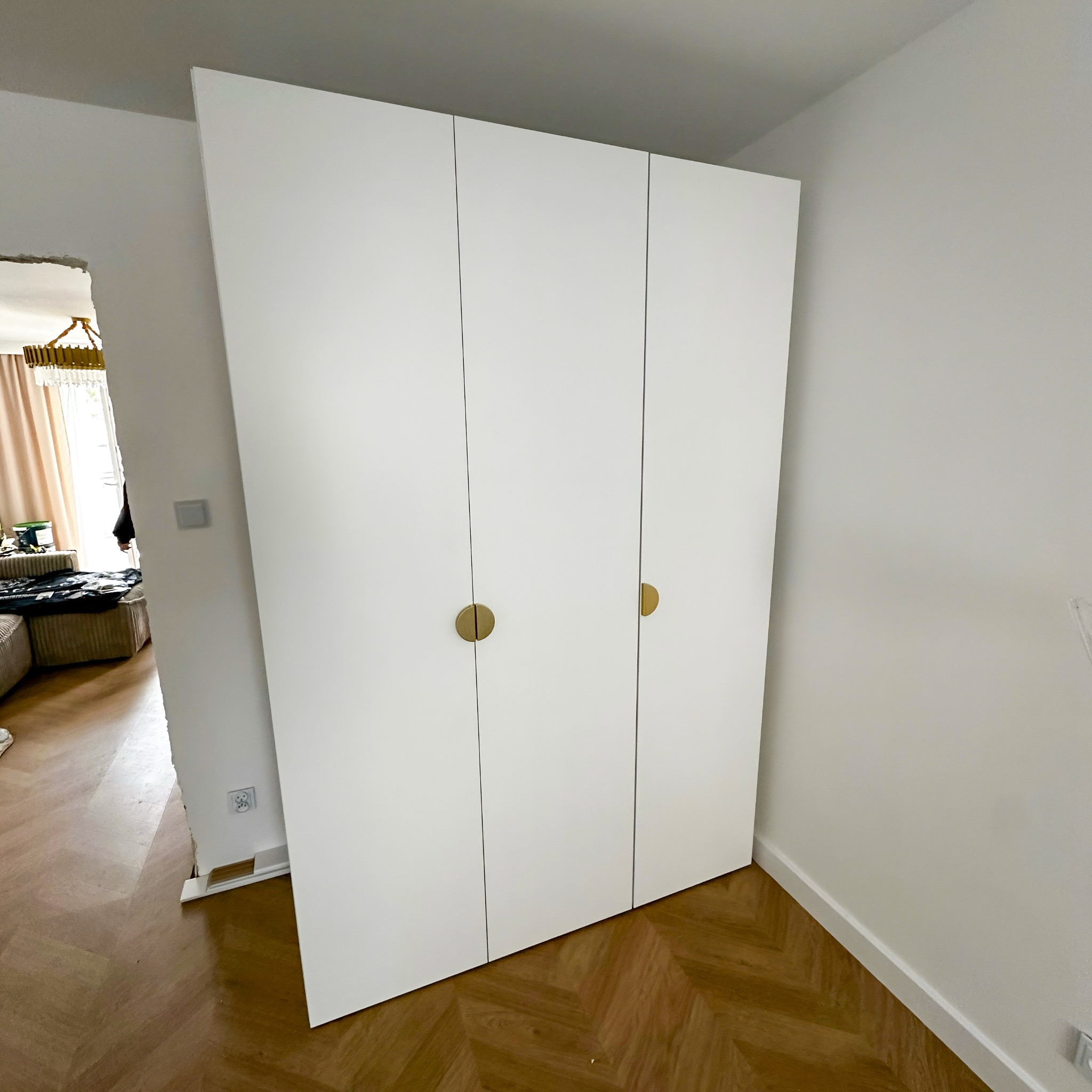 Biała szafa trzydrzwiowa z okrągłymi, złotymi uchwytami, ustawiona w rogu pokoju z parkietem w jodełkę. Widoczny fragment salonu z kanapą i żyrandolem.