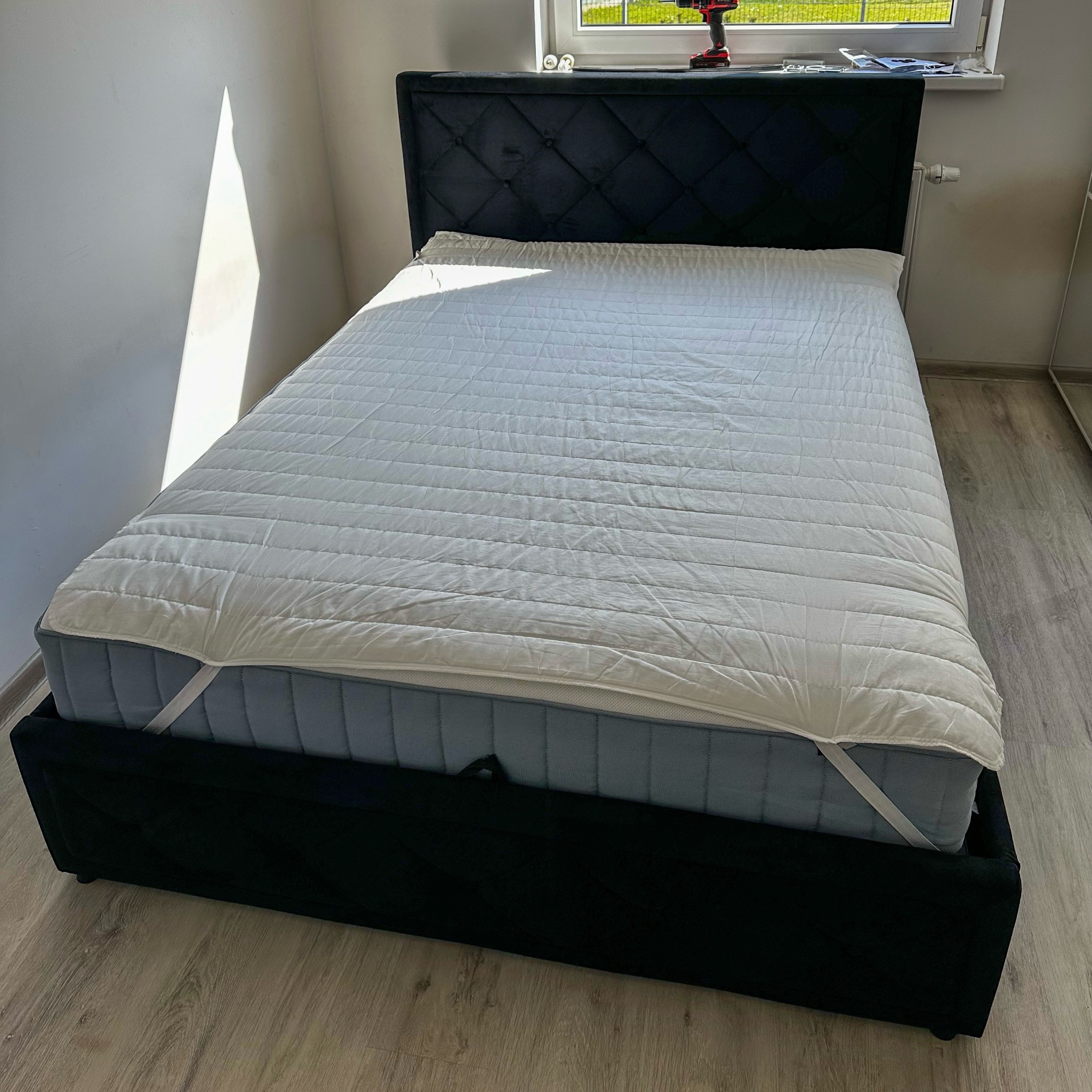 Czarne łóżko tapicerowane z pikowanym zagłówkiem, przy oknie w słonecznym pokoju. Na zagłówku widać narzędzia, sugerujące świeży montaż.