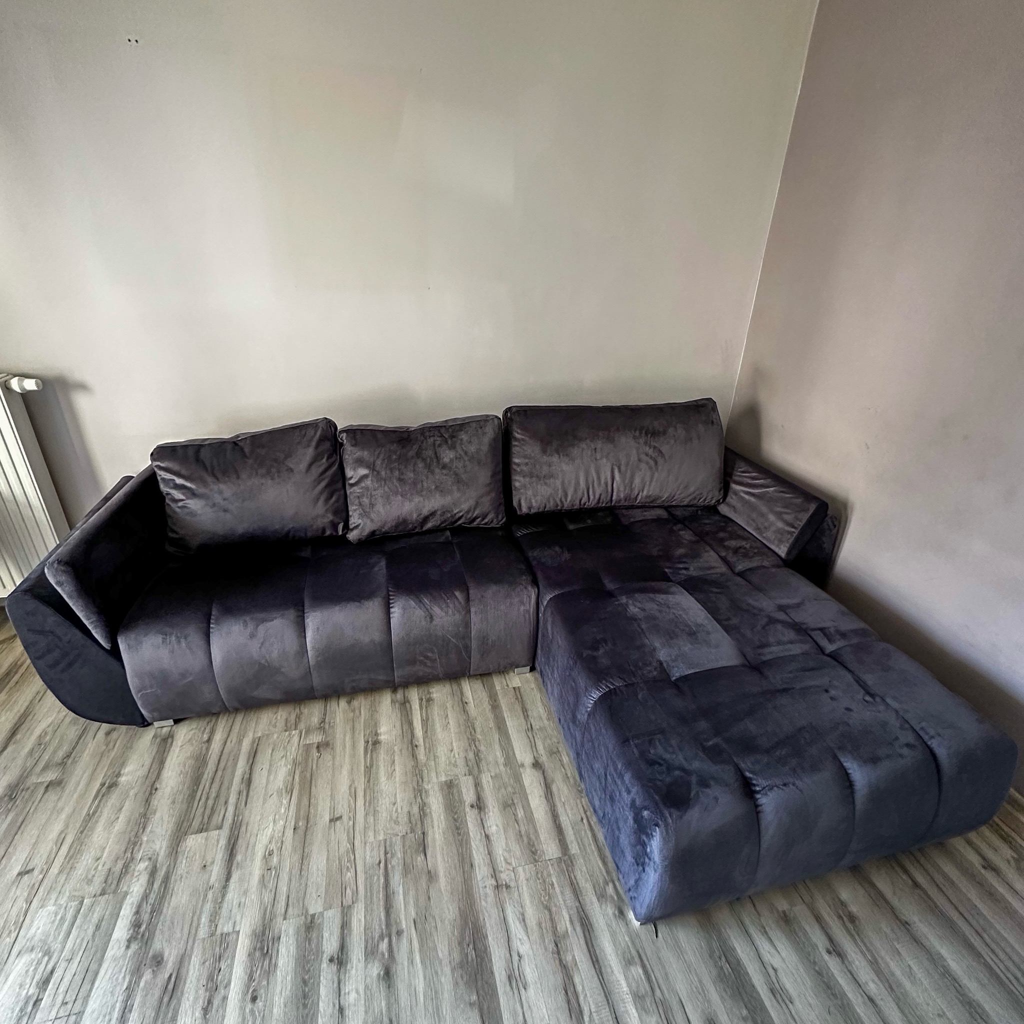 Ciemnoszara, welurowa sofa narożna z trzema poduszkami, ustawiona na panelach podłogowych w odcieniach szarości. Widok z przodu, oświetlenie naturalne.