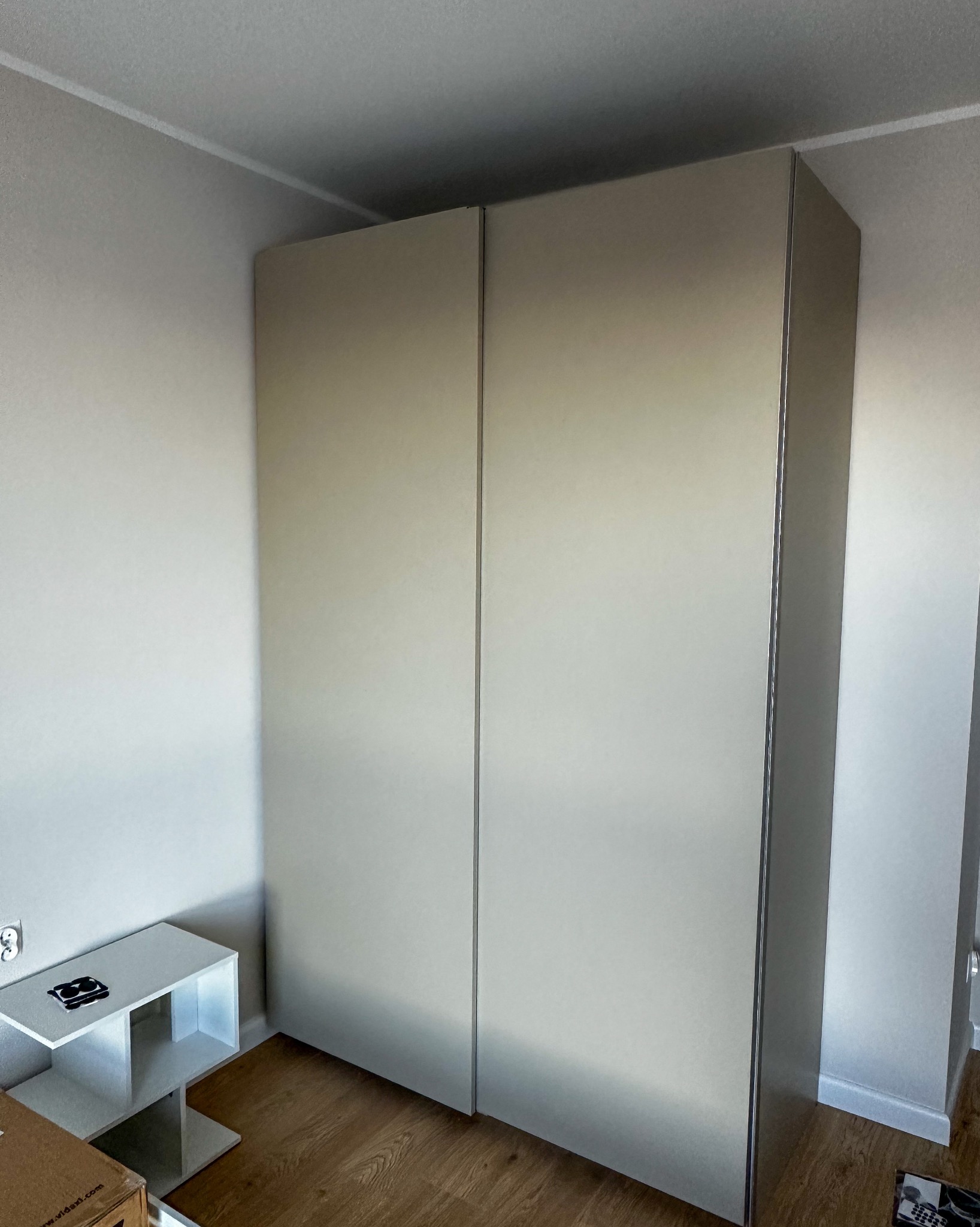 Beżowa szafa przesuwna z minimalistycznym wykończeniem, ustawiona w rogu pokoju z drewnianą podłogą i białą ścianą. Widoczny fragment stolika.