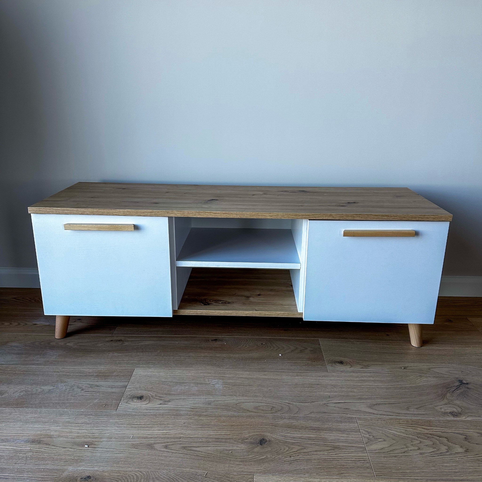 Biała szafka RTV z drewnianym blatem i uchwytami na tle szarej ściany i drewnianej podłogi. Widoczne nóżki w stylu skandynawskim.