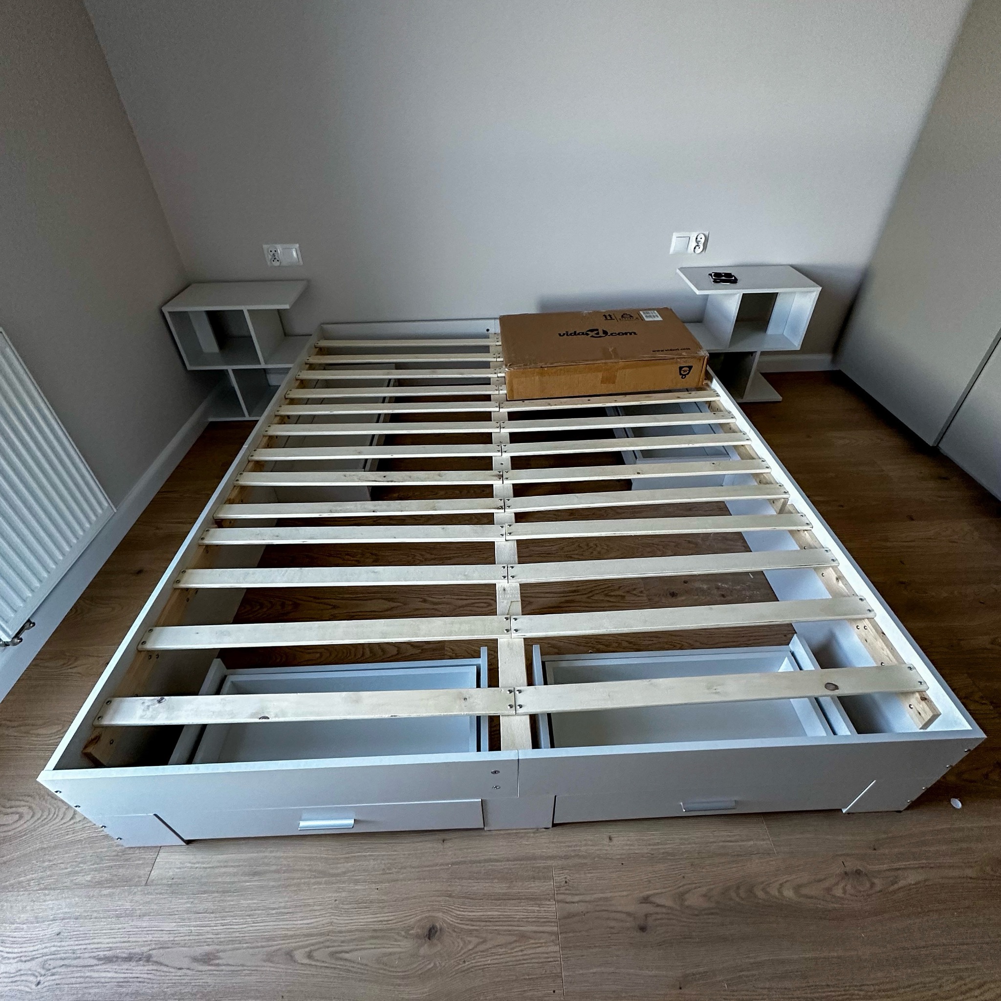 Biała rama łóżka z szufladami i stelażem listwowym, częściowo zmontowana, z pudełkiem na stelażu. Dwie białe szafki nocne po bokach. Minimalistyczne wnętrze.