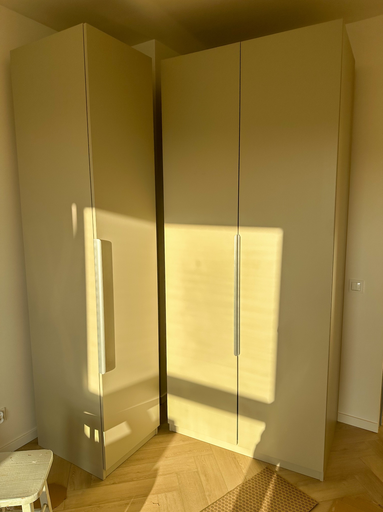 Dwie jasne szafy narożne z metalowymi uchwytami, oświetlone popołudniowym słońcem, stoją na parkiecie w pomieszczeniu. Widoczny fragment krzesła i dywanik.