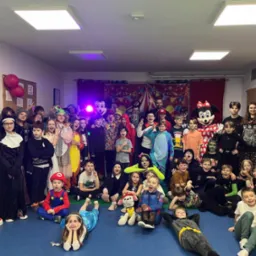 Grupowe zdjęcie dzieci w kostiumach karnawałowych, w tym Mario, Elsa i Batman, pozujących na niebieskiej podłodze w sali z dekoracjami cyrkowymi w tle.