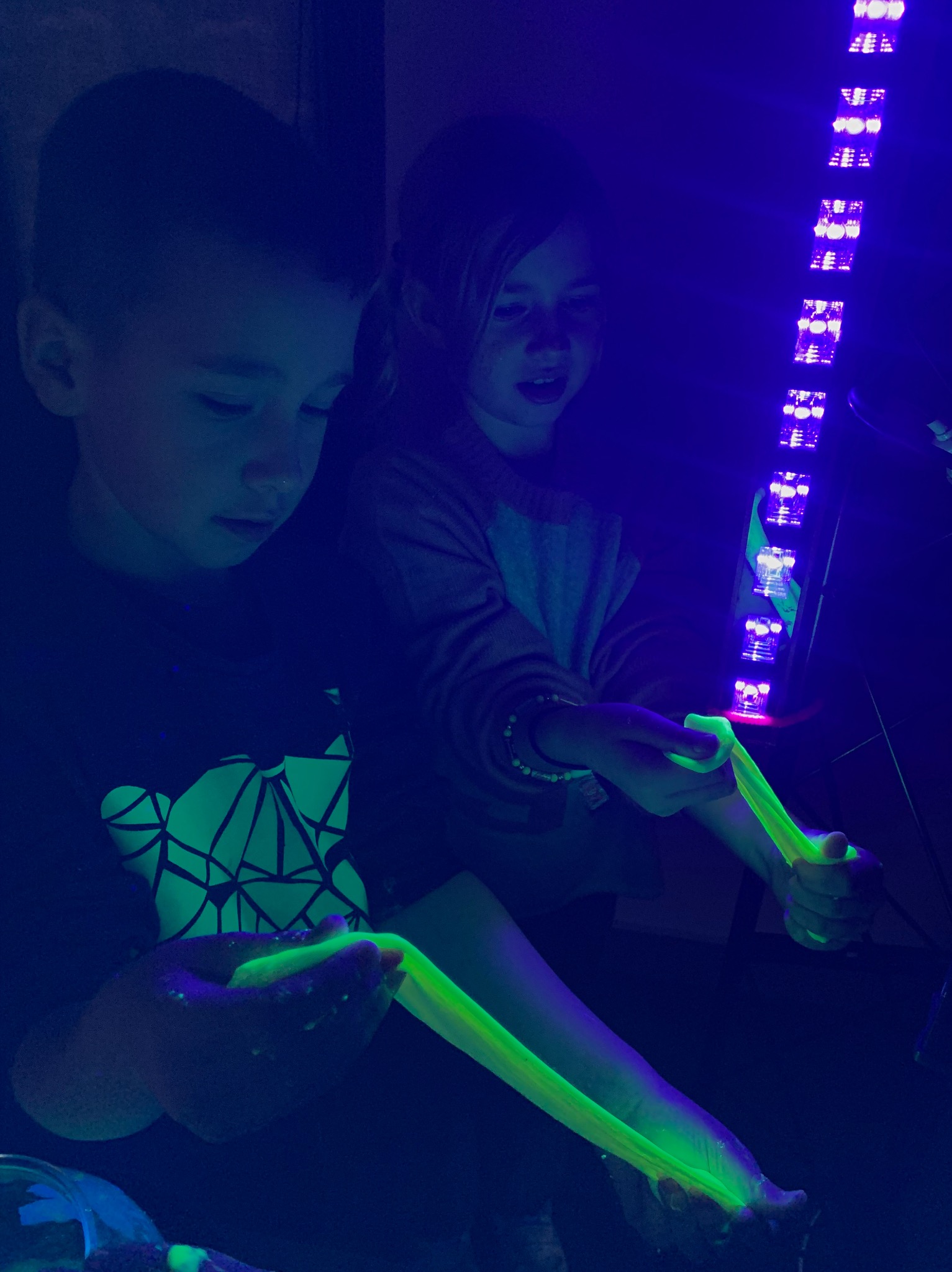 Dzieci bawią się świecącym slime w ciemności pod światłem UV. Chłopiec w koszulce z geometrycznym wzorem, dziewczynka z zaciekawieniem obserwuje zabawę.