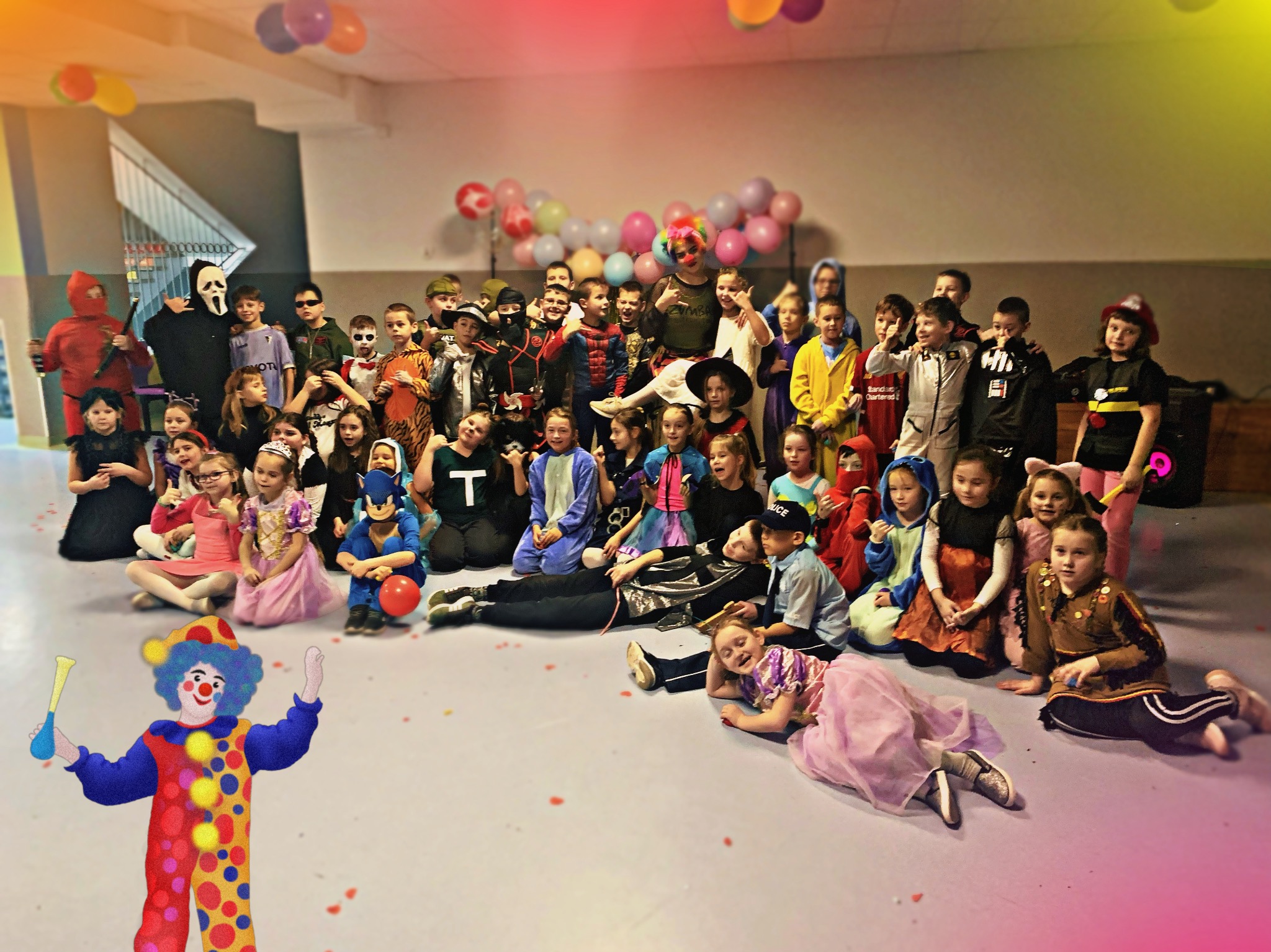 Duża grupa dzieci w kostiumach na imprezie, z animatorem-klaunem z przodu. Balony i dekoracje w tle. Różnorodne stroje, od superbohaterów po postacie z bajek.