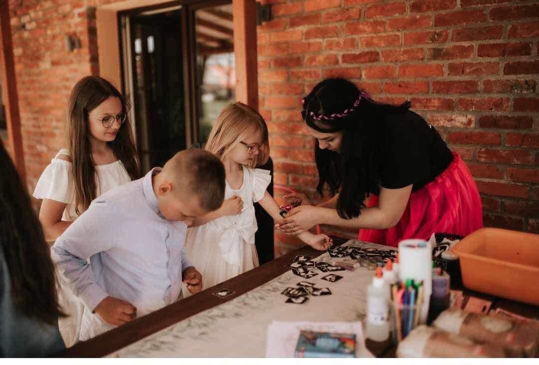 Animator maluje tatuaż na ramieniu dziecka podczas przyjęcia. Dzieci obserwują z zaciekawieniem. Na stole farby i szablony. Ceglana ściana w tle.