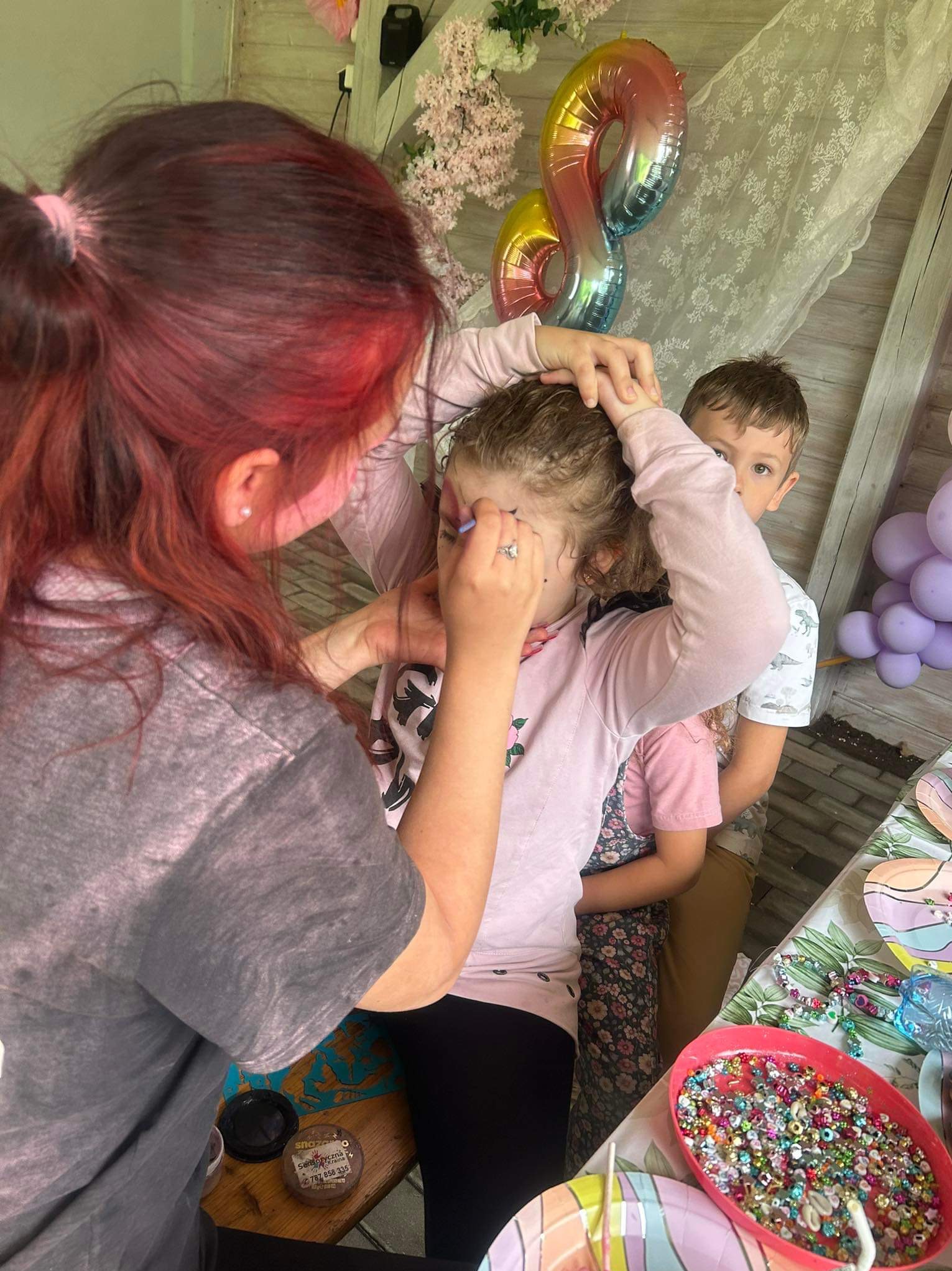 Animator maluje twarz dziewczynce na urodzinach, w tle balony w kształcie cyfry osiem i dwoje dzieci. Na stole akcesoria do malowania i koraliki.