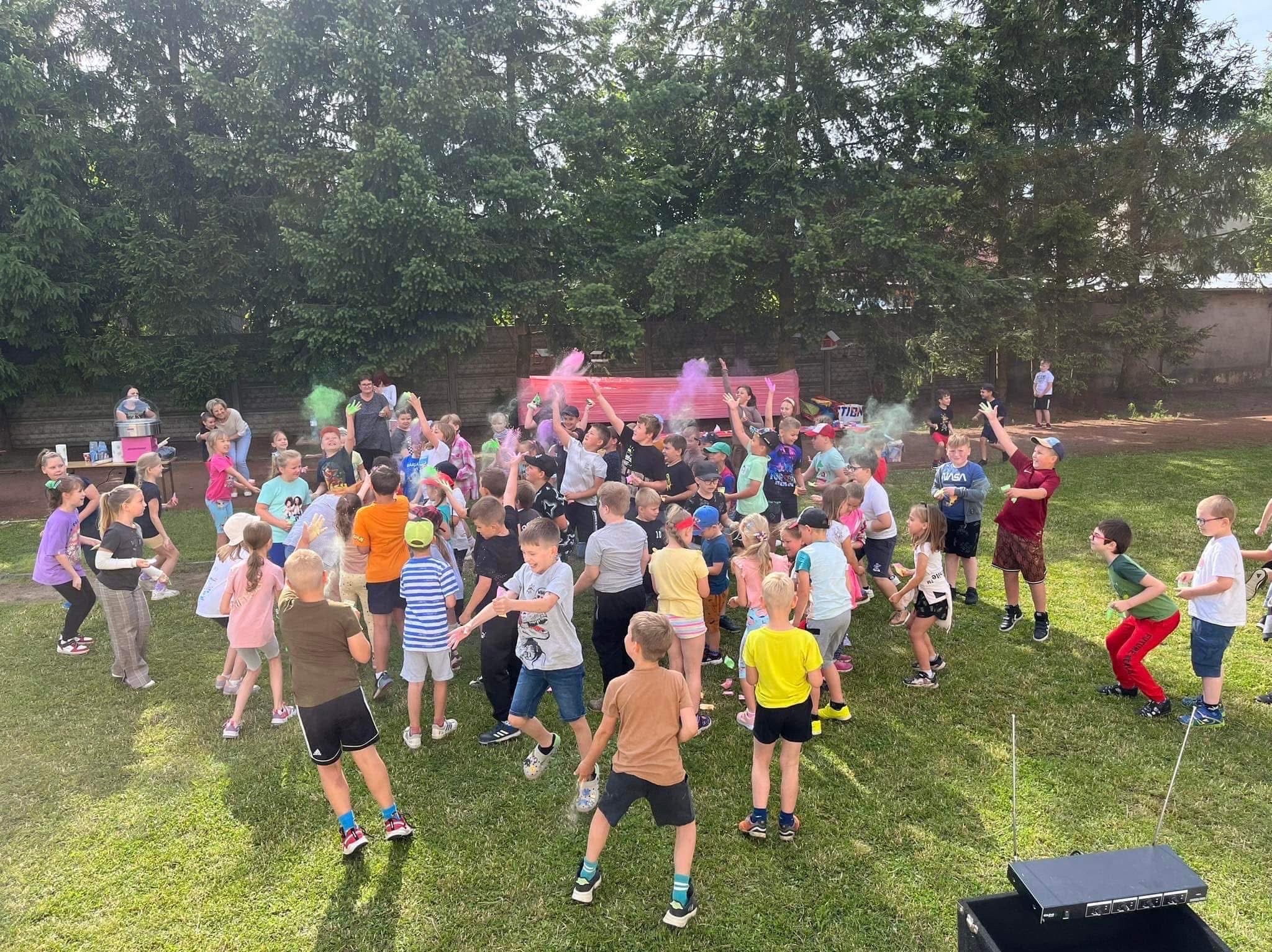 Grupa dzieci rzuca kolorowymi proszkami holi podczas zabawy na zielonej trawie. W tle drzewa i budynki. Dzieci w różnym wieku, w kolorowych ubraniach.