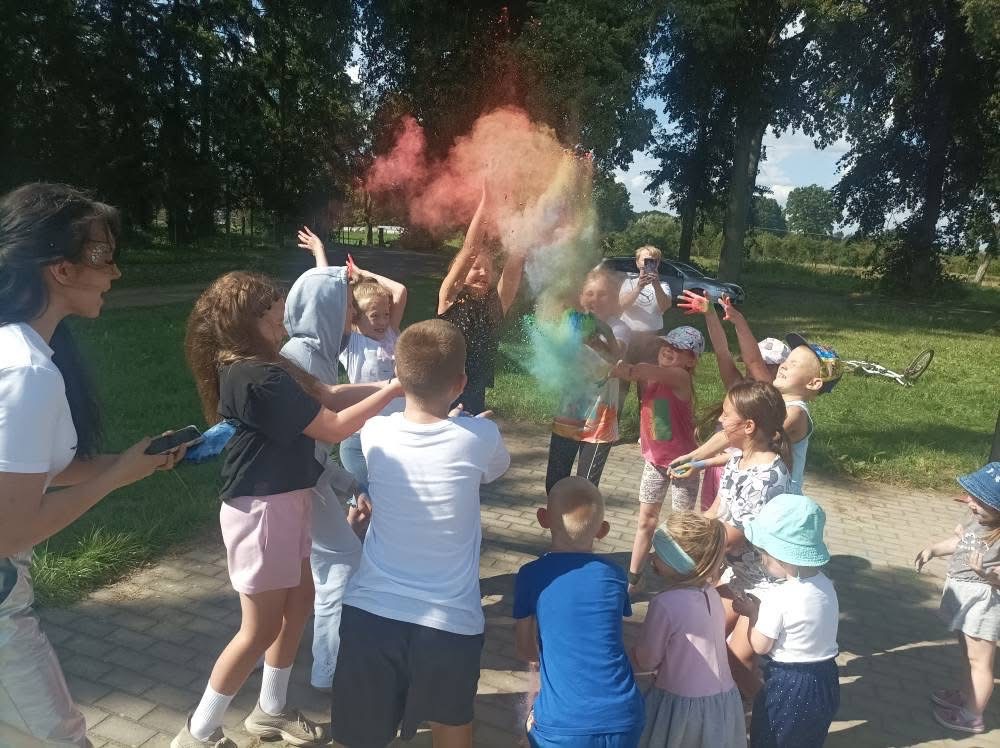 Grupa dzieci rzuca kolorowe proszki holi podczas zabawy na zewnątrz, w tle drzewa i osoba robiąca zdjęcia telefonem. Kolorowy dym w powietrzu.