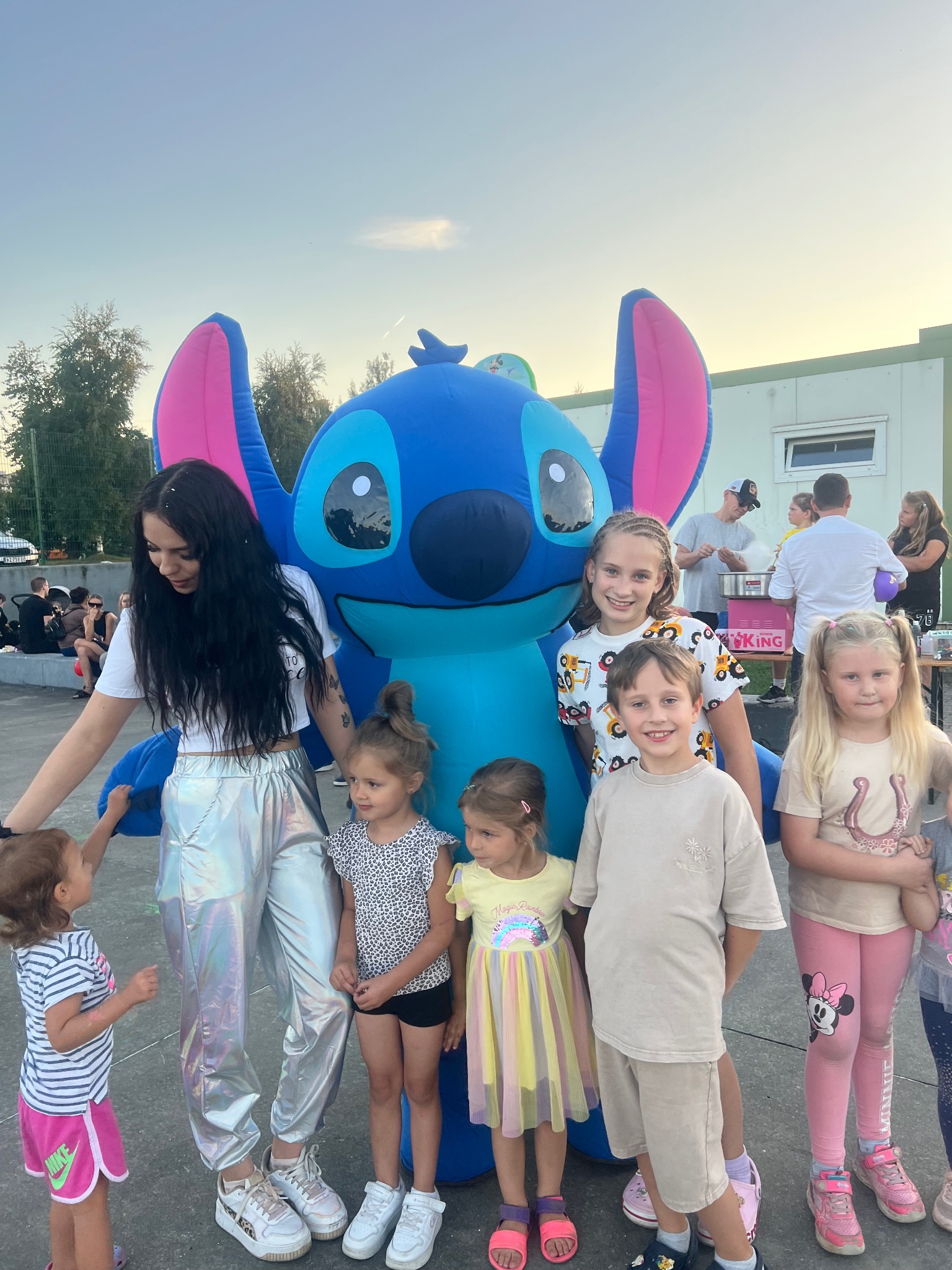Animator w srebrnych spodniach i grupa dzieci pozują obok dmuchanej postaci Stitch na tle plenerowej imprezy z watą cukrową.