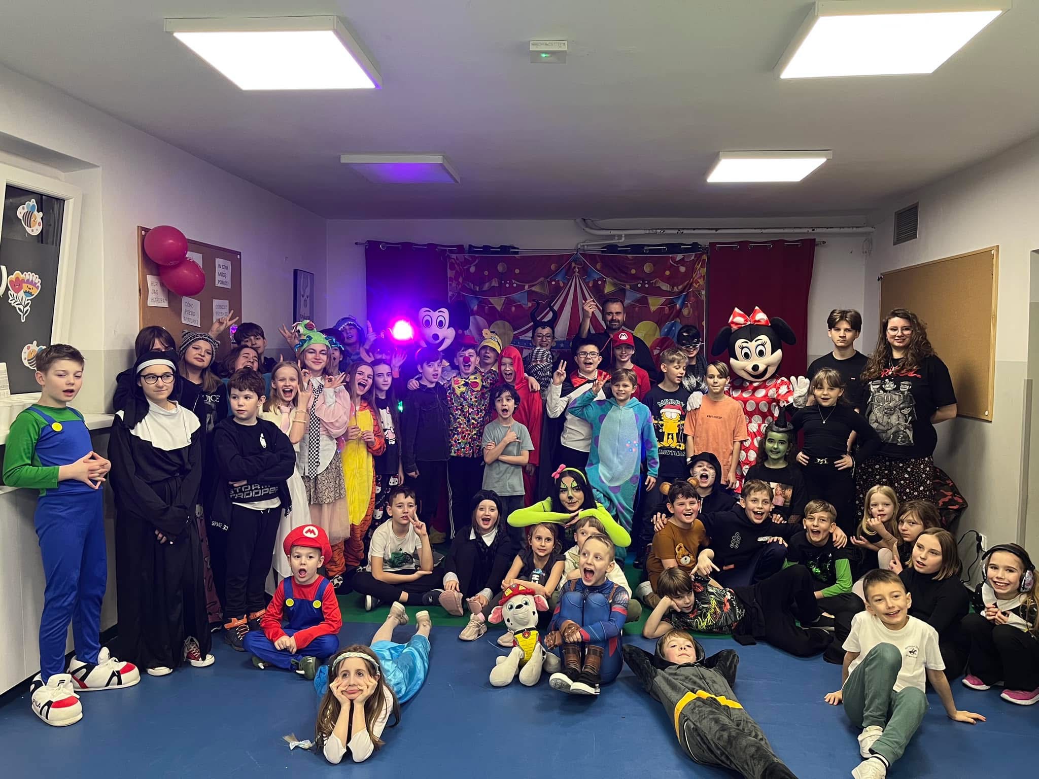 Grupowe zdjęcie dzieci w kostiumach karnawałowych, w tym Mario, Elsa i Batman, pozujących na niebieskiej podłodze w sali z dekoracjami cyrkowymi w tle.