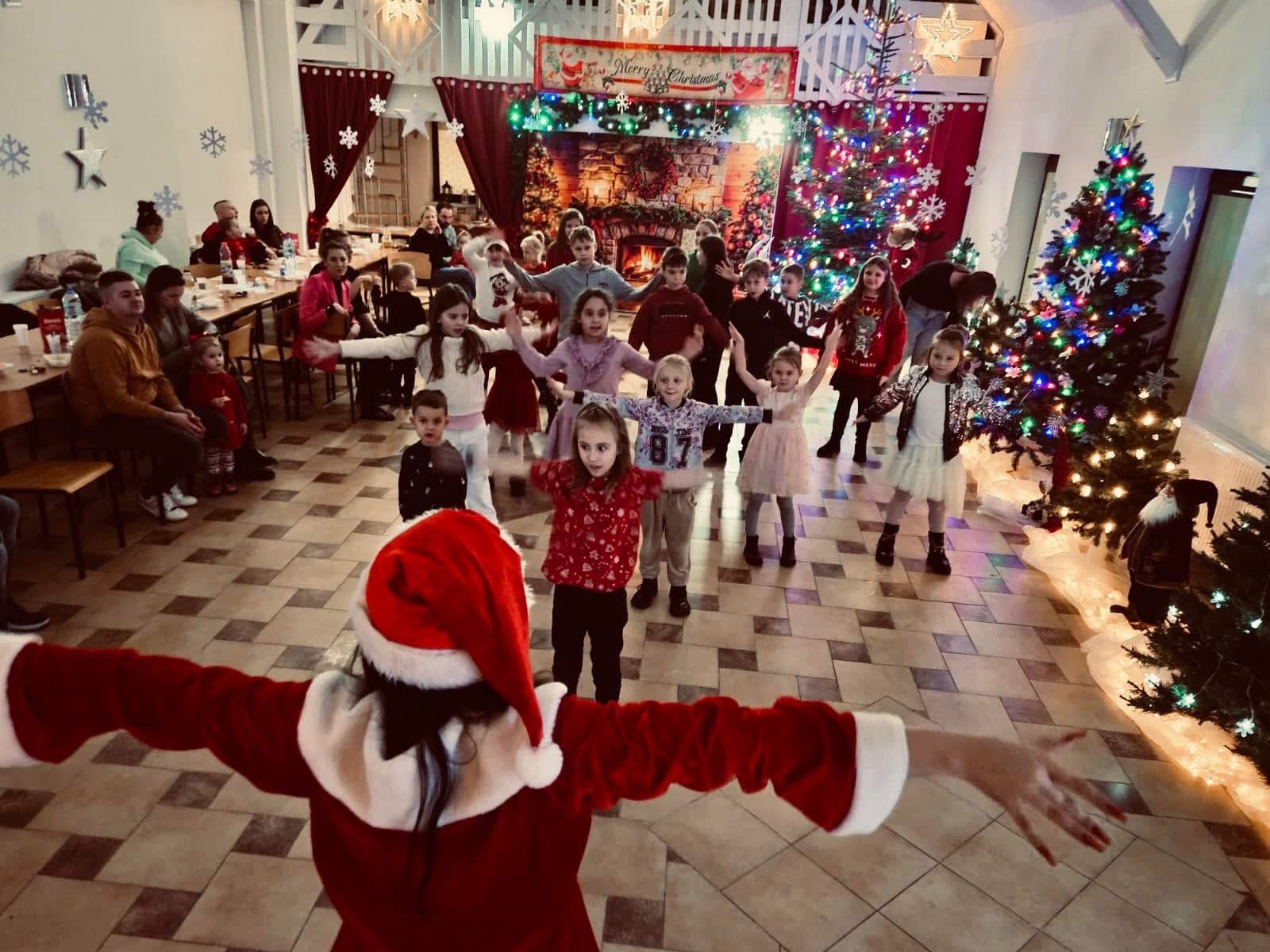 Animator w stroju Mikołaja prowadzi grupę dzieci podczas świątecznej zabawy w udekorowanym pomieszczeniu z choinkami i dekoracjami bożonarodzeniowymi.