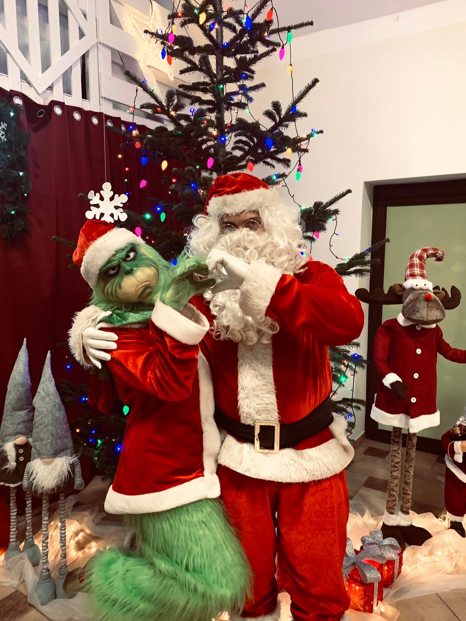 Świąteczna sceneria: Grinch obejmuje Świętego Mikołaja na tle choinki z kolorowymi lampkami, tworząc radosną atmosferę zabawy i świątecznego klimatu.