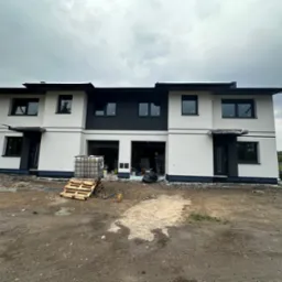 Dwukondygnacyjny bliźniak z białą elewacją i czarnymi akcentami pod pochmurnym niebem. Na pierwszym planie teren budowy z materiałami i paletami.