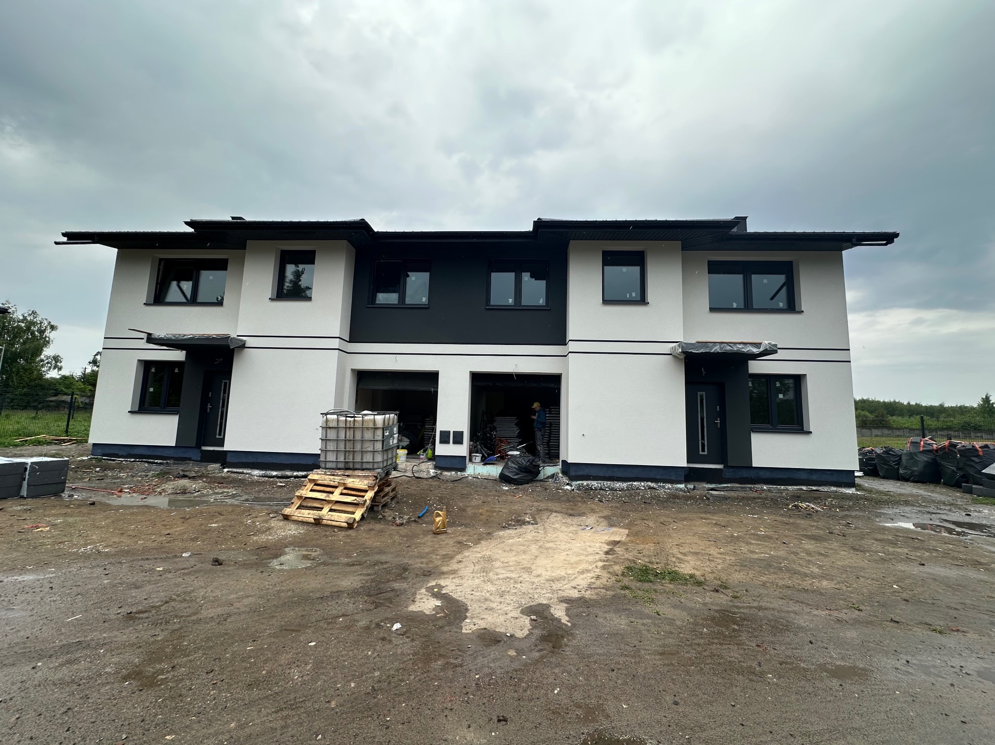 Dwukondygnacyjny bliźniak z białą elewacją i czarnymi akcentami pod pochmurnym niebem. Na pierwszym planie teren budowy z materiałami i paletami.