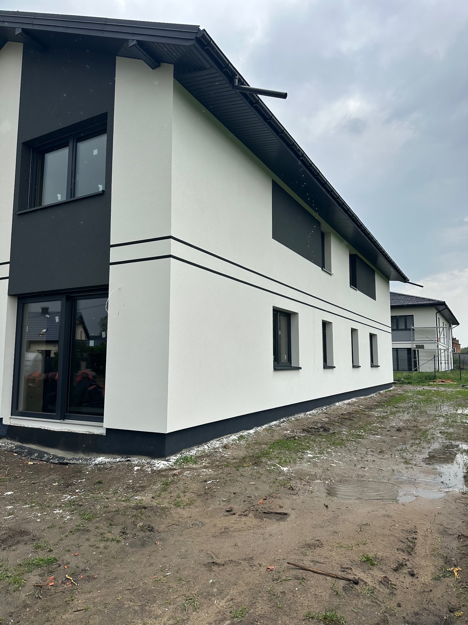 Dwukondygnacyjny dom z białą elewacją i czarnymi akcentami, widoczny z boku, z mokrą ziemią w tle i drugim budynkiem w oddali. Nowoczesny design.