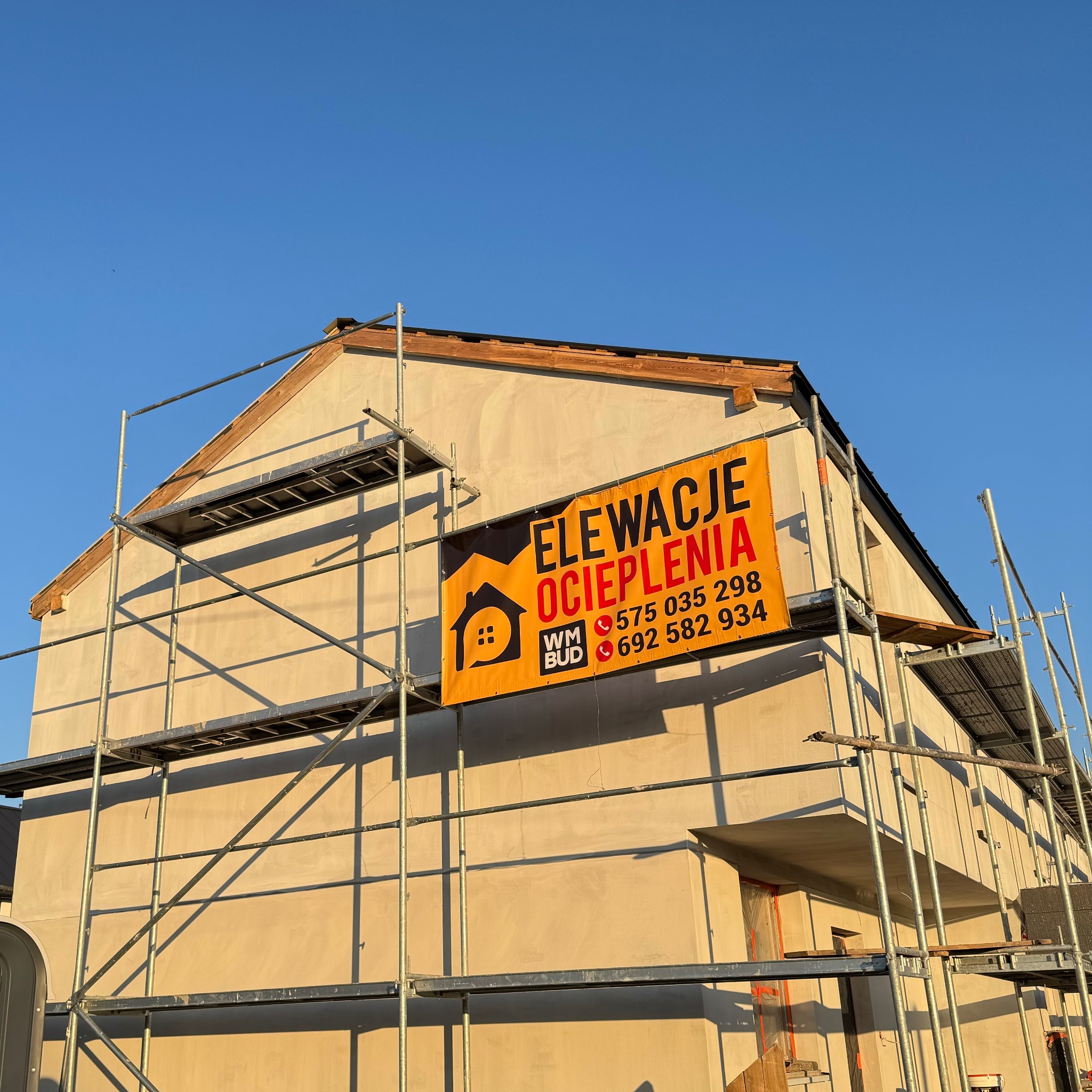Dom w trakcie ocieplania, z widocznym rusztowaniem i banerem reklamowym firmy oferującej elewacje i ocieplenia. Częściowo otynkowana elewacja w kolorze beżowym.