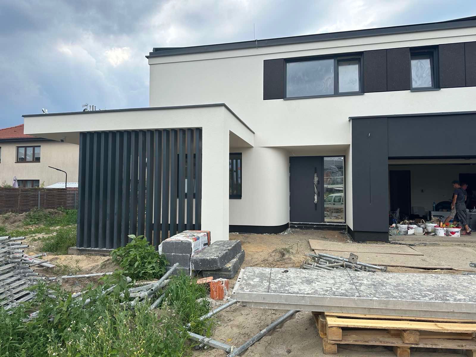 Nowoczesny dom z białą elewacją i grafitowymi akcentami, widoczne rusztowania i materiały budowlane. Minimalistyczny design, surowe linie, garaż z ażurową przesłoną.