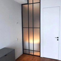 Integralstal Serhii Bondarenko - Nowoczesna, wewnętrzna zabudowa z metalową ramą i matowym szkłem, dzieląca przestrzeń obok białych drzwi w minimalistycznym wnętrzu z drewnianą podłogą.