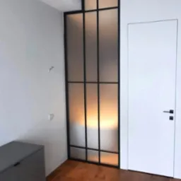Nowoczesna, wewnętrzna zabudowa z metalową ramą i matowym szkłem, dzieląca przestrzeń obok białych drzwi w minimalistycznym wnętrzu z drewnianą podłogą.