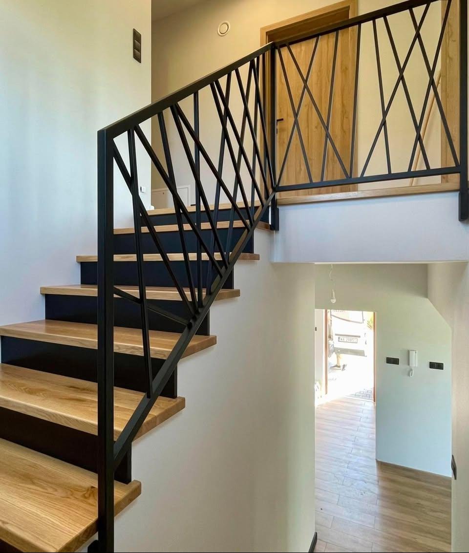 Nowoczesne schody z drewnianymi stopniami i czarną, geometryczną balustradą. Widok z dołu, jasne wnętrze domu, drewniana podłoga w tle.