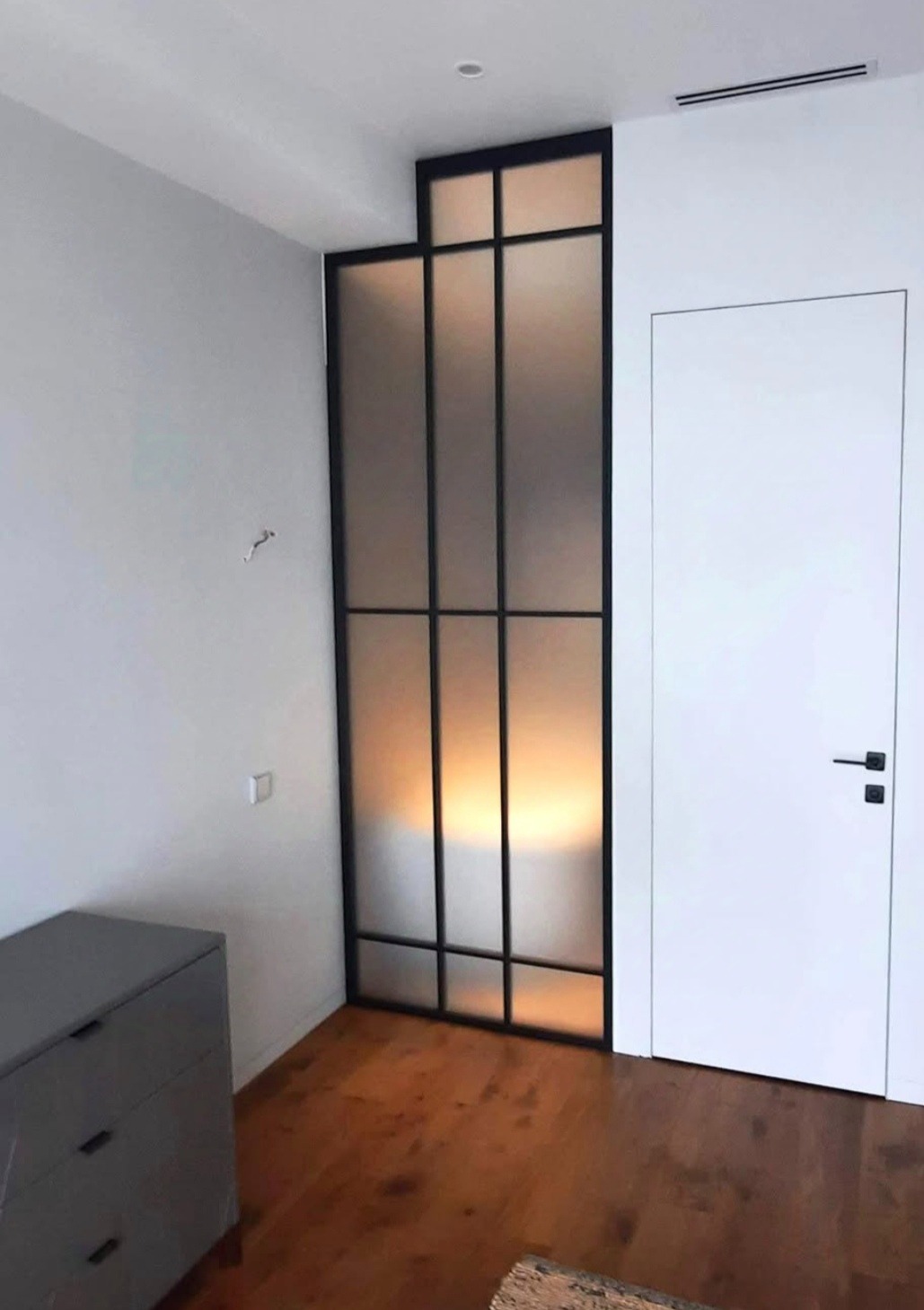 Nowoczesna, wewnętrzna zabudowa z metalową ramą i matowym szkłem, dzieląca przestrzeń obok białych drzwi w minimalistycznym wnętrzu z drewnianą podłogą.