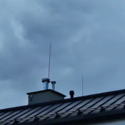 Dwa kominy wentylacyjne ze stali nierdzewnej na dachu budynku z metalową dachówką, widoczne na tle pochmurnego nieba. Dodatkowo widoczny odgromnik.