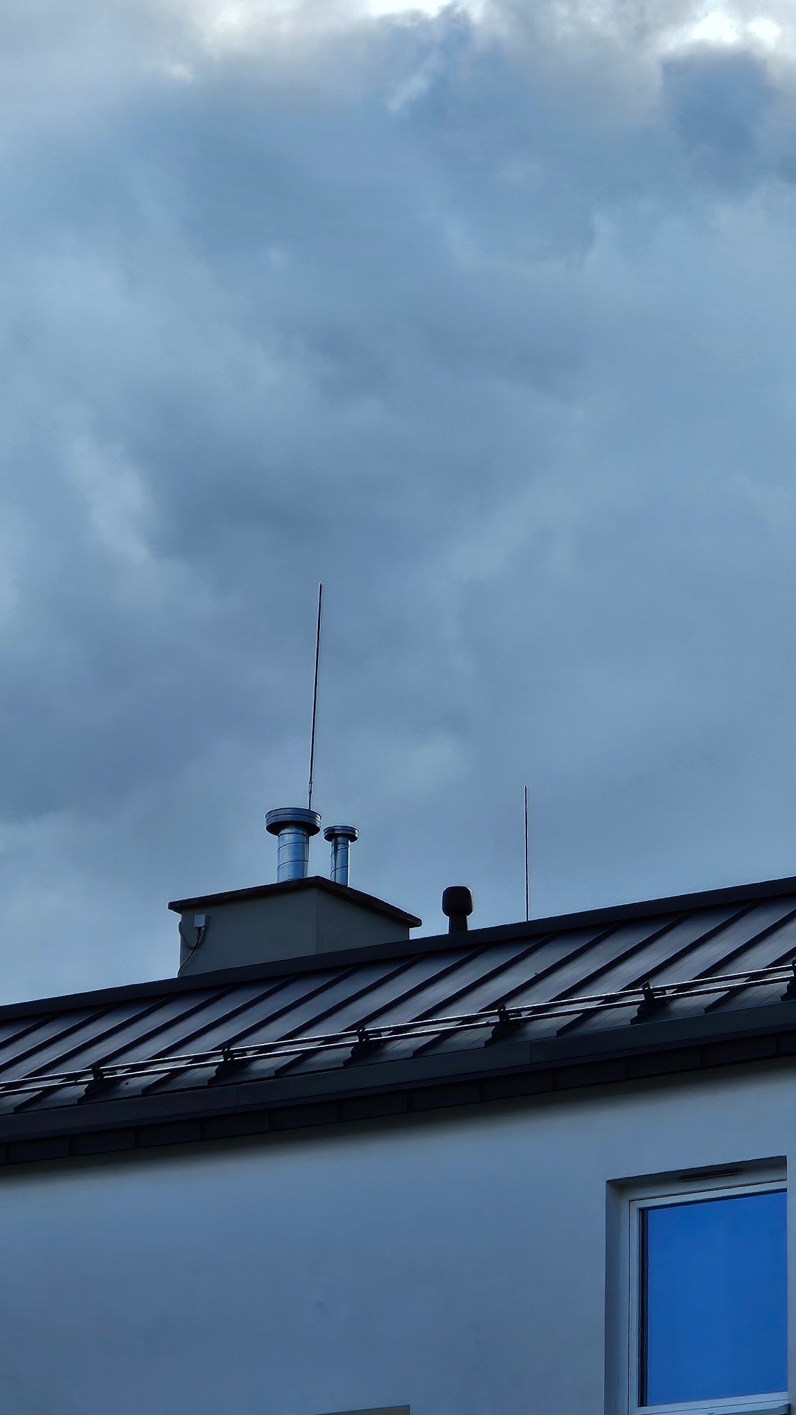 Dwa kominy wentylacyjne ze stali nierdzewnej na dachu budynku z metalową dachówką, widoczne na tle pochmurnego nieba. Dodatkowo widoczny odgromnik.