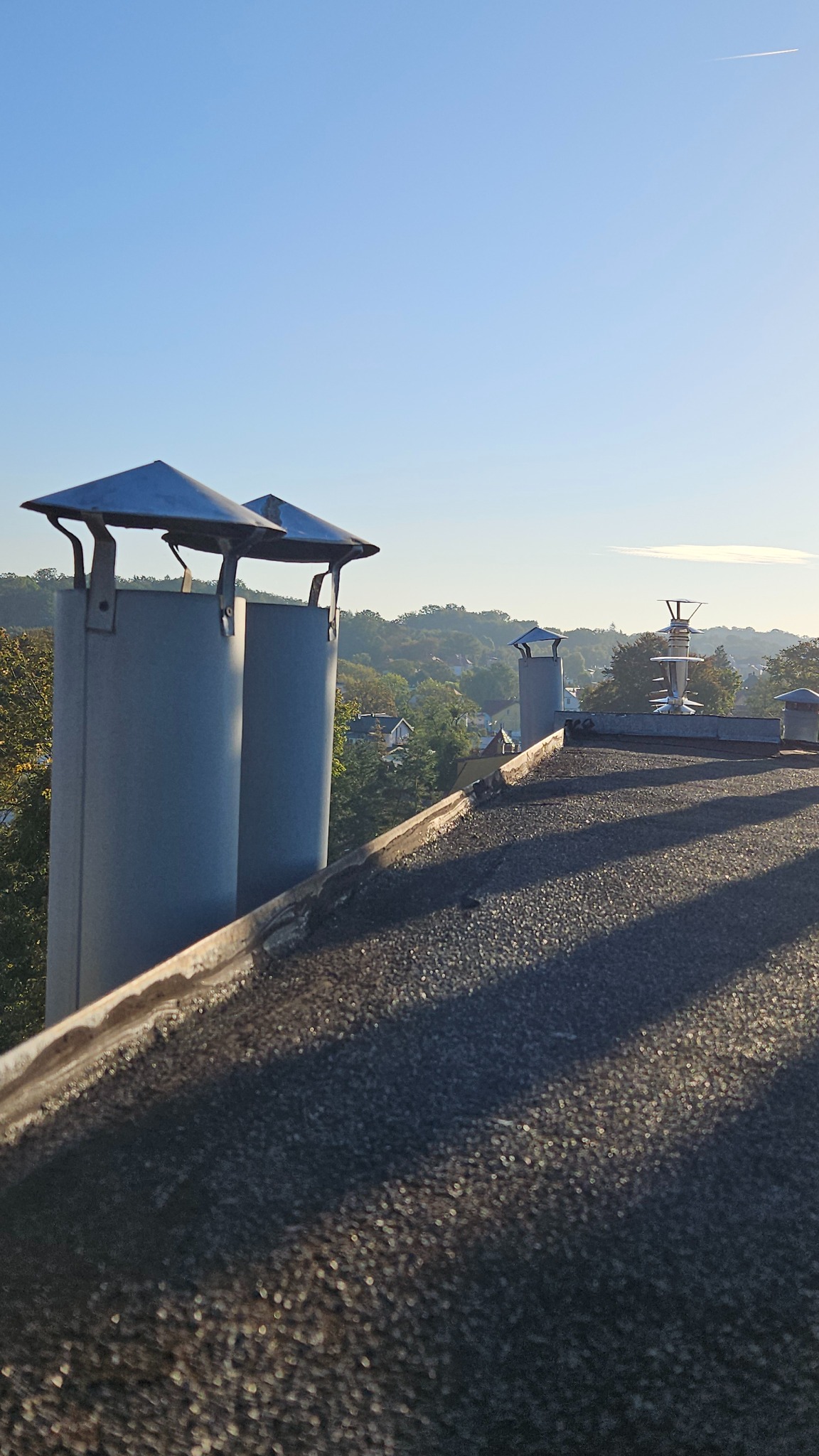 Dwa metalowe kominy z daszkami na bitumicznym dachu, w tle zieleń drzew i zabudowania, błękitne niebo. Ujęcie z dołu, podkreślające wysokość konstrukcji.