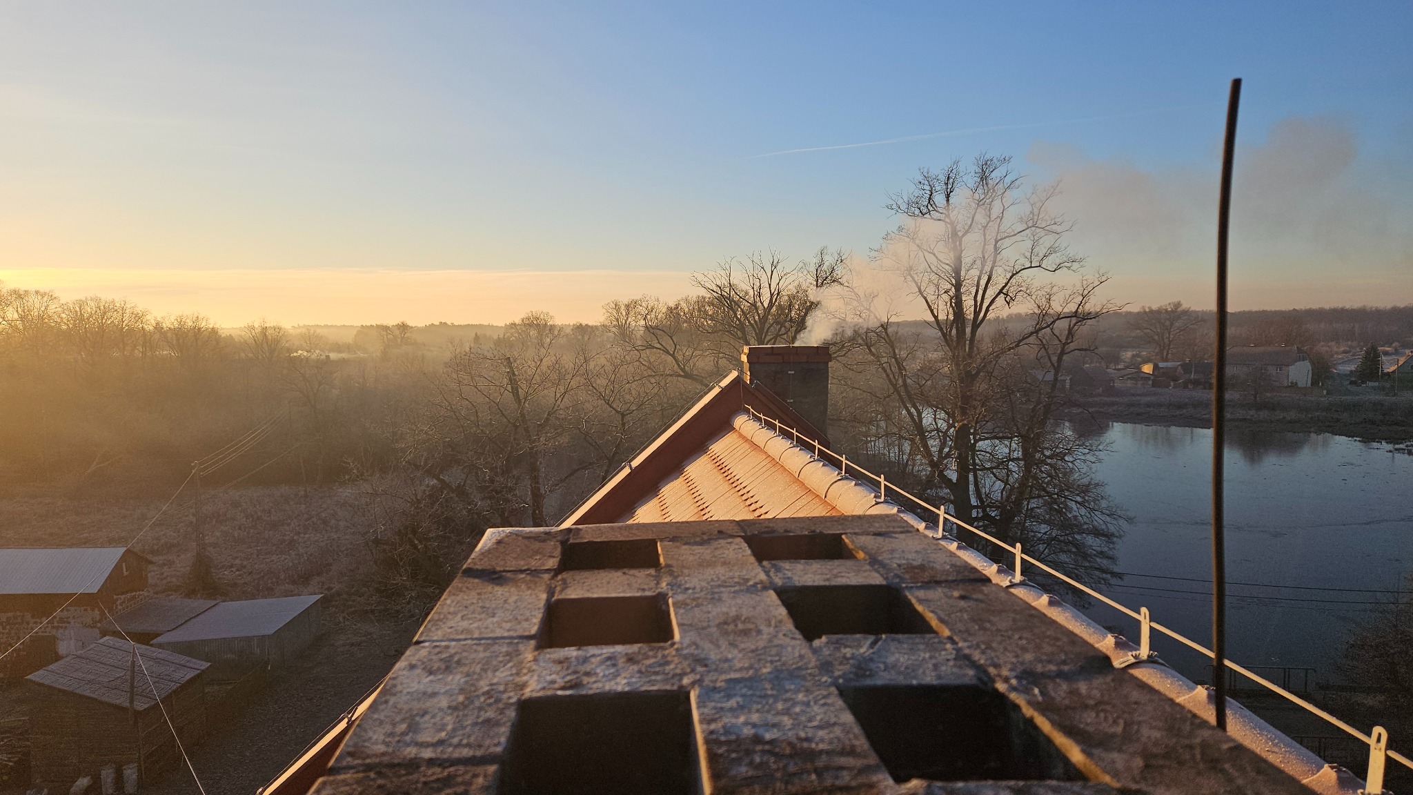 Komin z cegły z widokiem na zalesiony krajobraz w zimowej aurze. Z komina unosi się dym, co sugeruje jego użytkowanie.