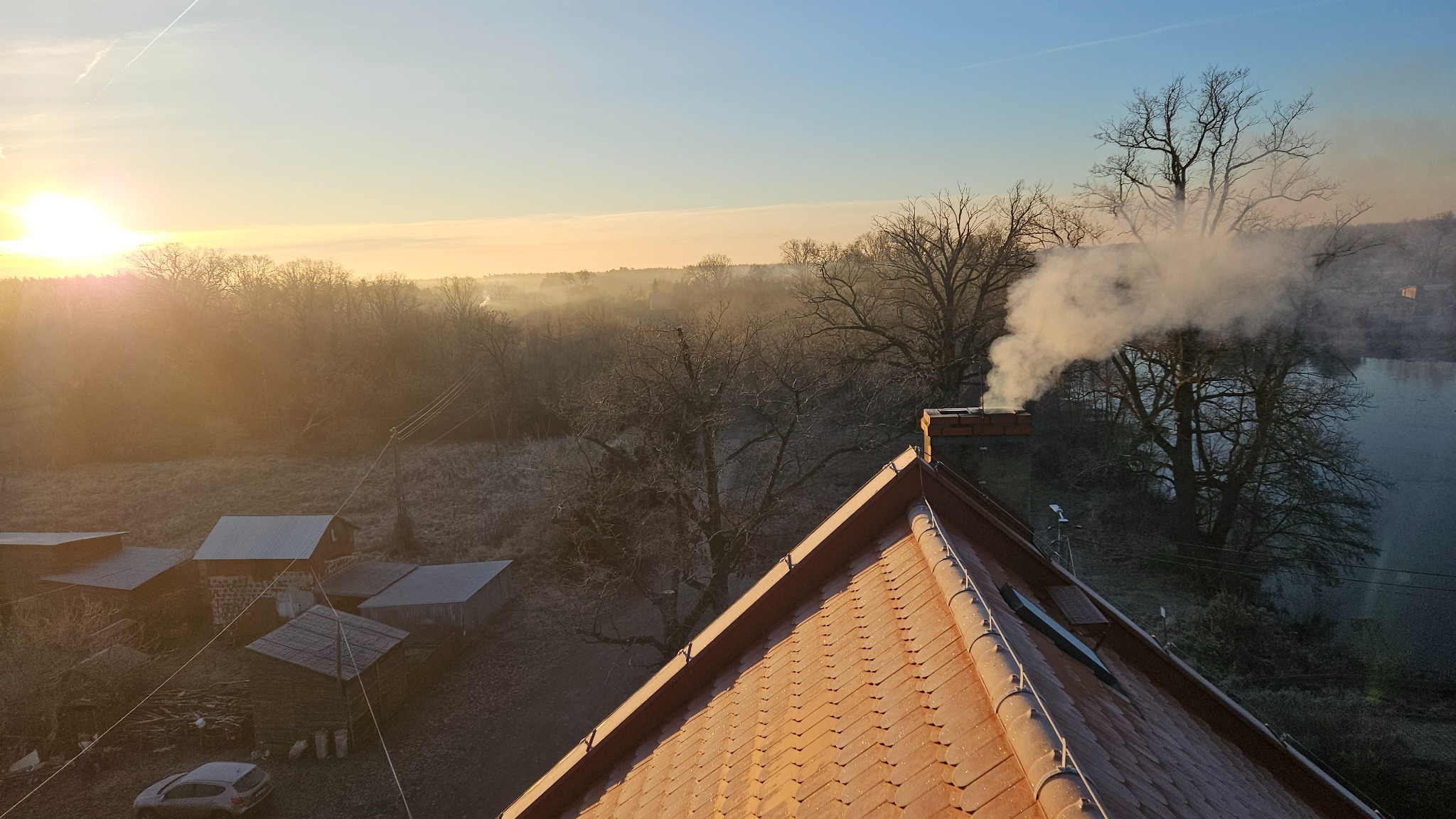 Dym wydobywa się z ceglanego komina na dachu domu o świcie, z widokiem na zalesiony krajobraz i wschodzące słońce. Widoczna część dachu pokryta czerwoną dachówką.
