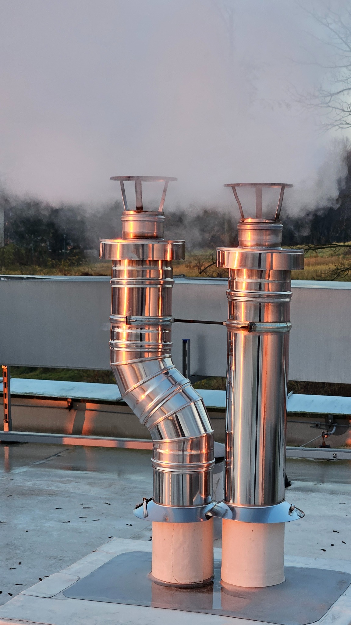 Dwa lśniące, metalowe kominy wentylacyjne na dachu, z widoczną parą/dymem wydobywającą się z jednego z nich, na tle drzew i nieba o zachodzie słońca.