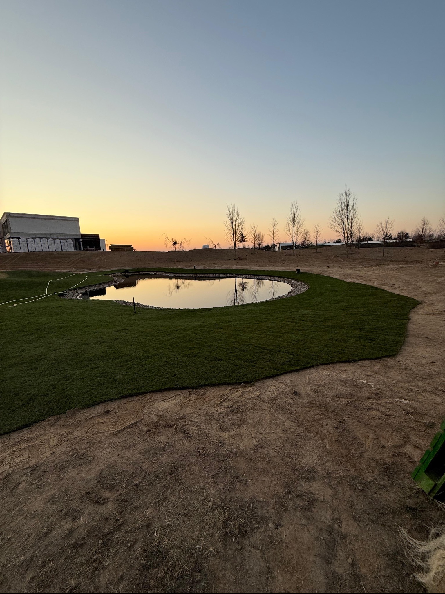 Świeżo ułożona trawa z rolki wokół oczka wodnego o zmierzchu. W tle budynek i drzewa bez liści, ziemia przygotowana pod dalsze prace.