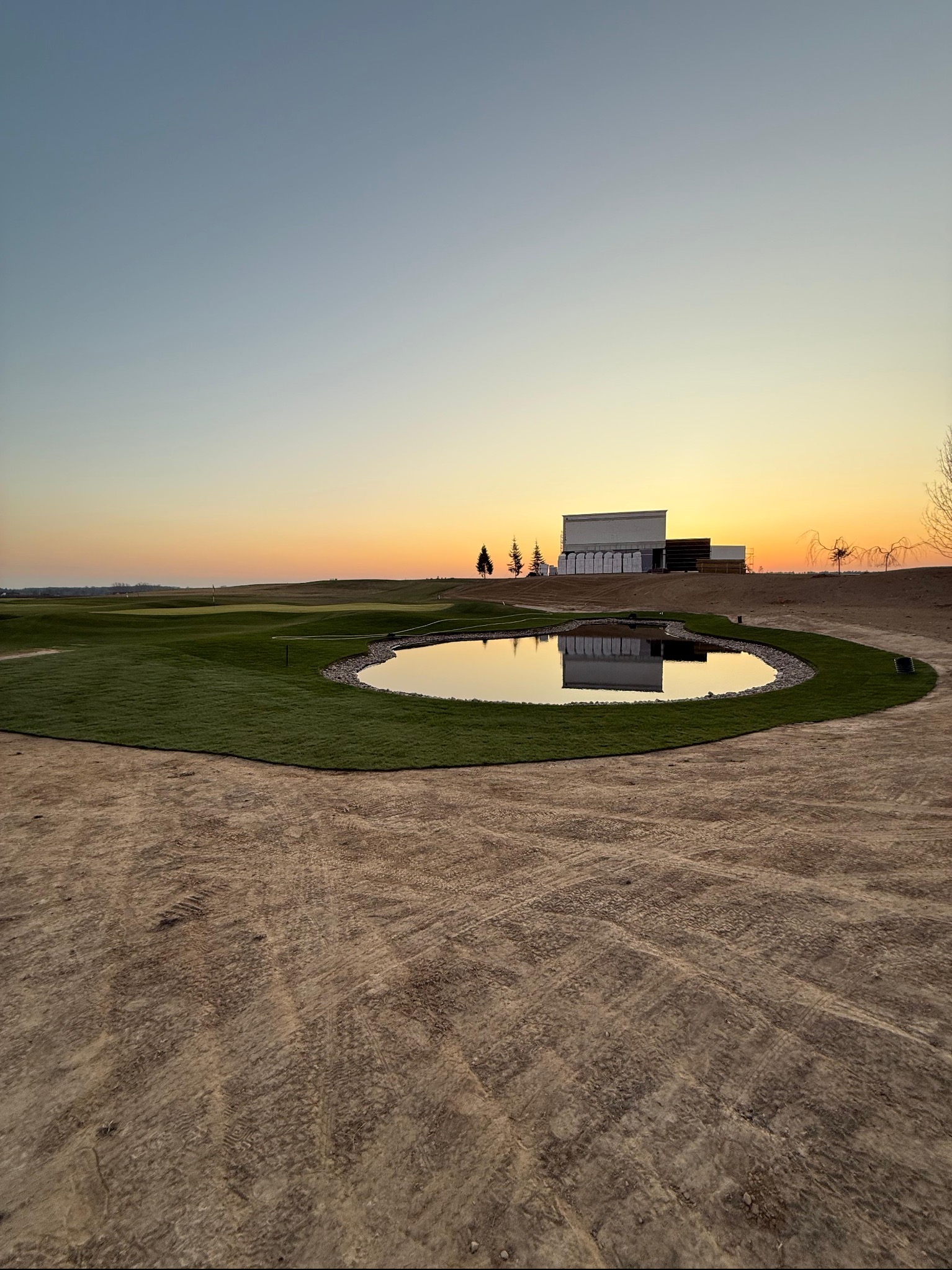 Krajobraz po niwelacji terenu z oczkiem wodnym i budynkiem w tle, oświetlony zachodzącym słońcem. Widoczne ślady opon na piasku.