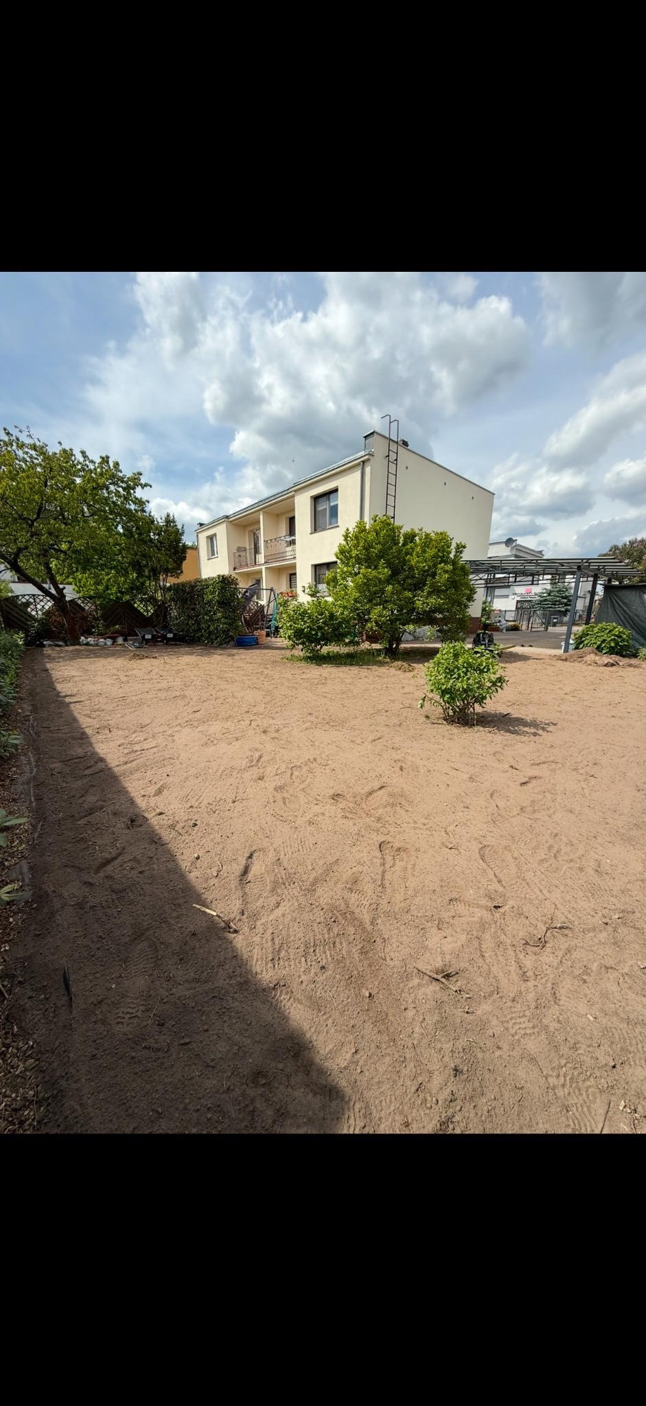Przygotowany teren pod trawnik z widocznymi śladami, w tle dom z balkonem i zielona roślinność, pochmurne niebo. Niwelacja terenu przed założeniem trawnika.
