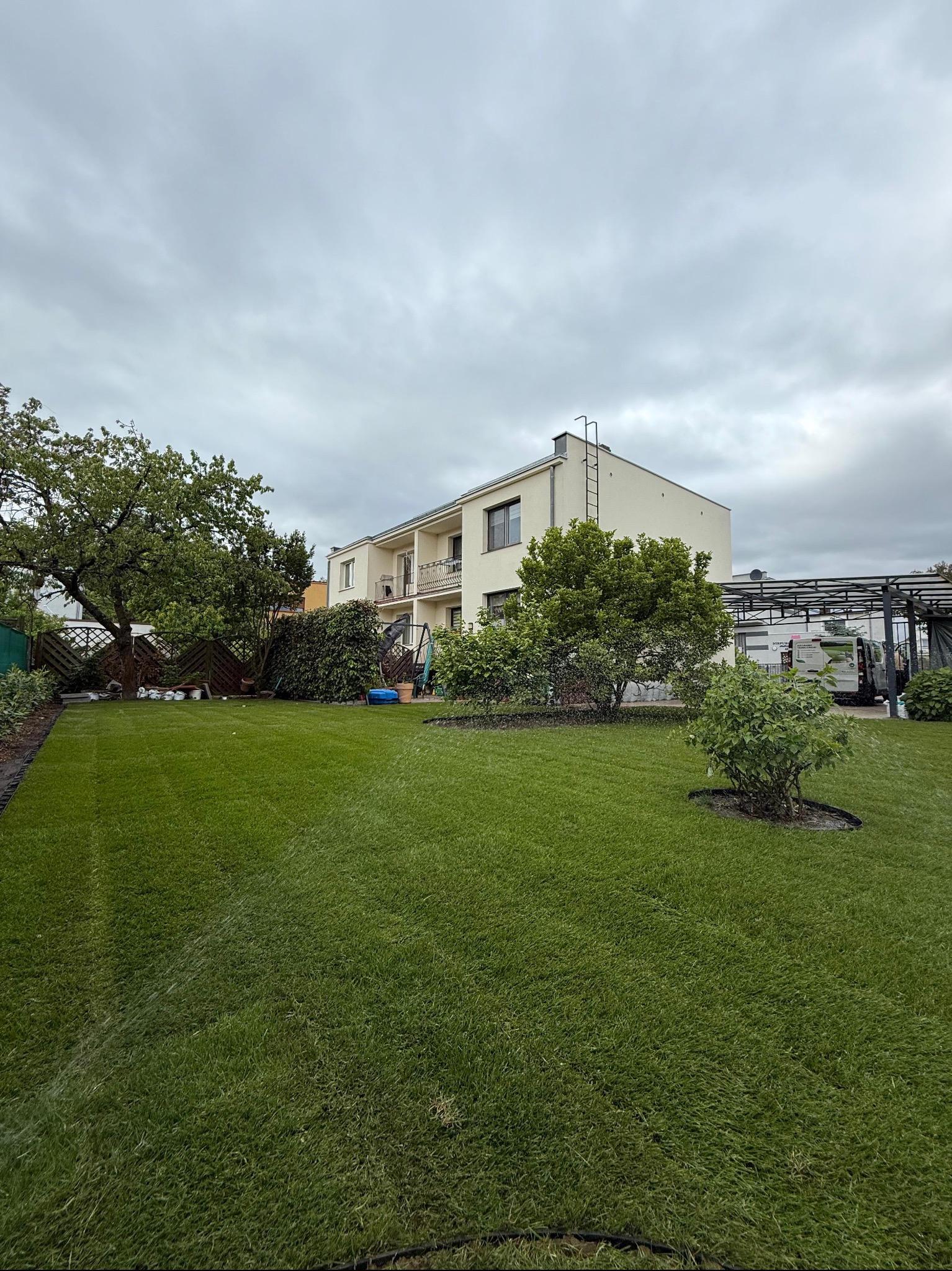 Świeżo ułożona, intensywnie zielona trawa z rolki przed nowoczesnym domem w pochmurny dzień, z widocznymi śladami podlewania i elementami ogrodu.