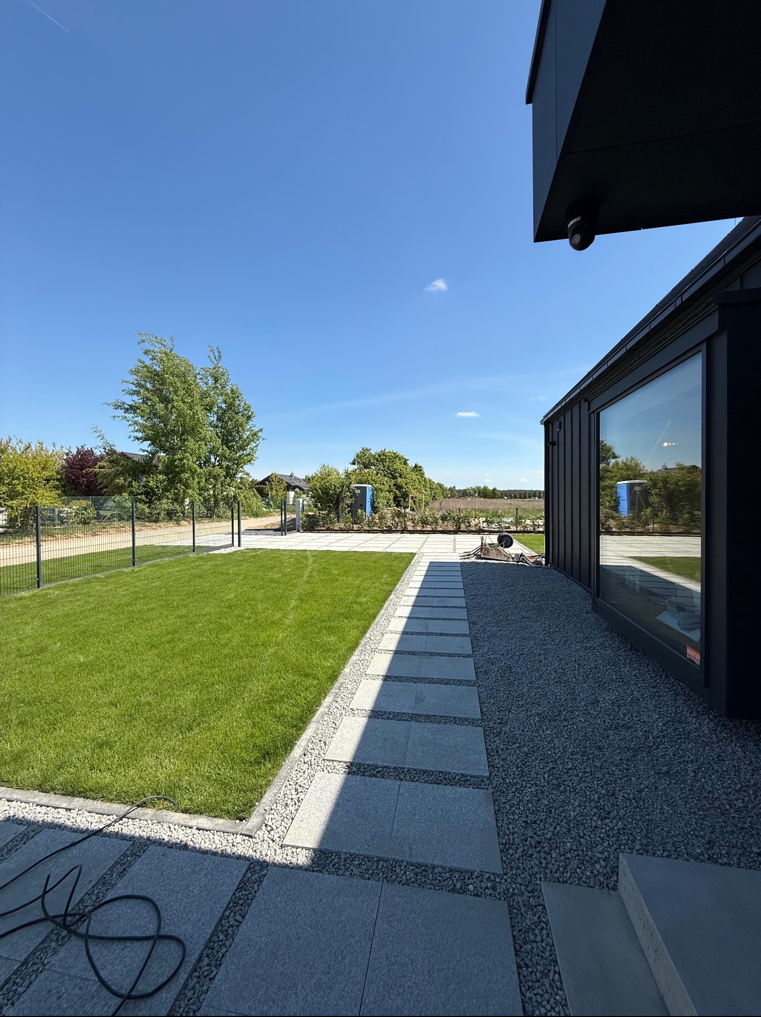Nowoczesny ogród z trawnikiem, ścieżką z płyt i szarego żwiru przy czarnym budynku. Widoczne drzewo i ogrodzenie.