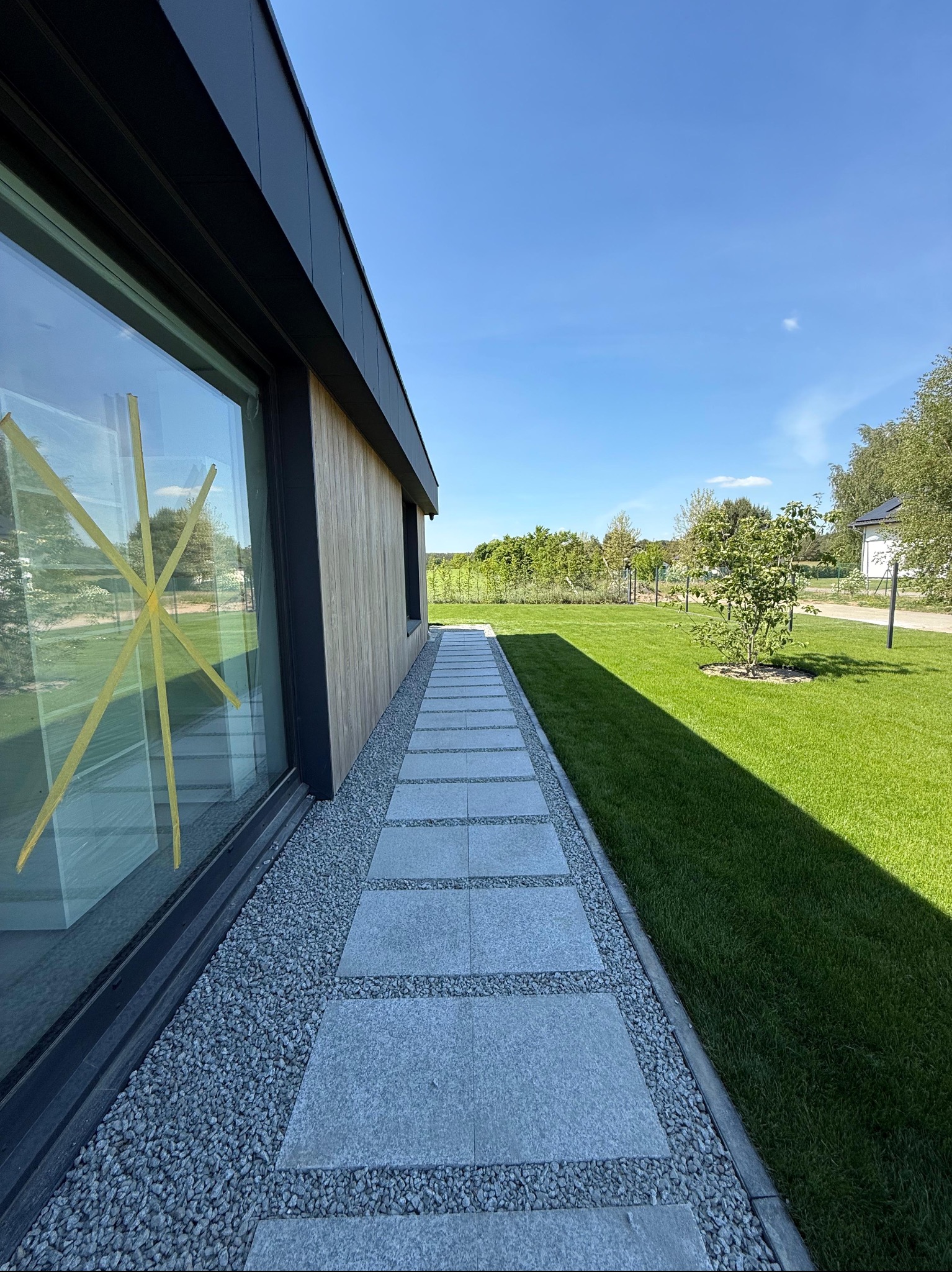 Nowoczesny ogród z równo przystrzyżoną trawą, kamienną ścieżką i minimalistycznym domem z dużym oknem zabezpieczonym taśmą. Słoneczny dzień.