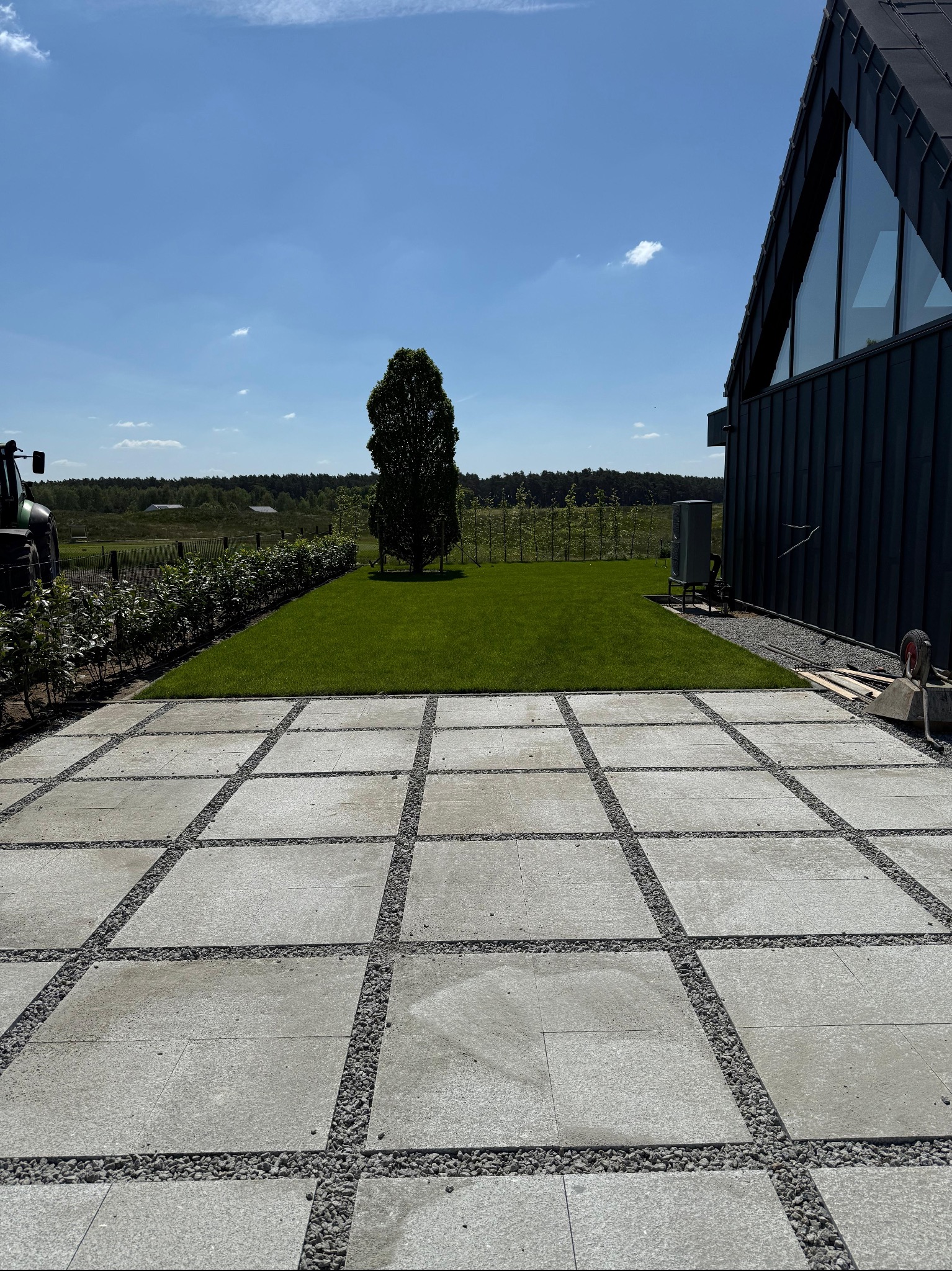 Nowoczesny ogród: trawnik z rolki, drzewo, kostka z kamieniami, czarny dom z dużymi oknami i traktor w tle, w słoneczny dzień.