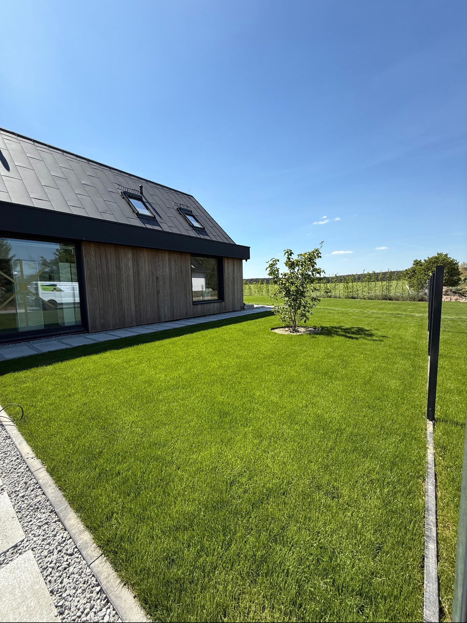 Nowoczesny dom z drewnianą elewacją i idealnie zielonym trawnikiem. W tle widoczny żywopłot i błękitne niebo. Perfekcyjnie utrzymany ogród przydomowy.