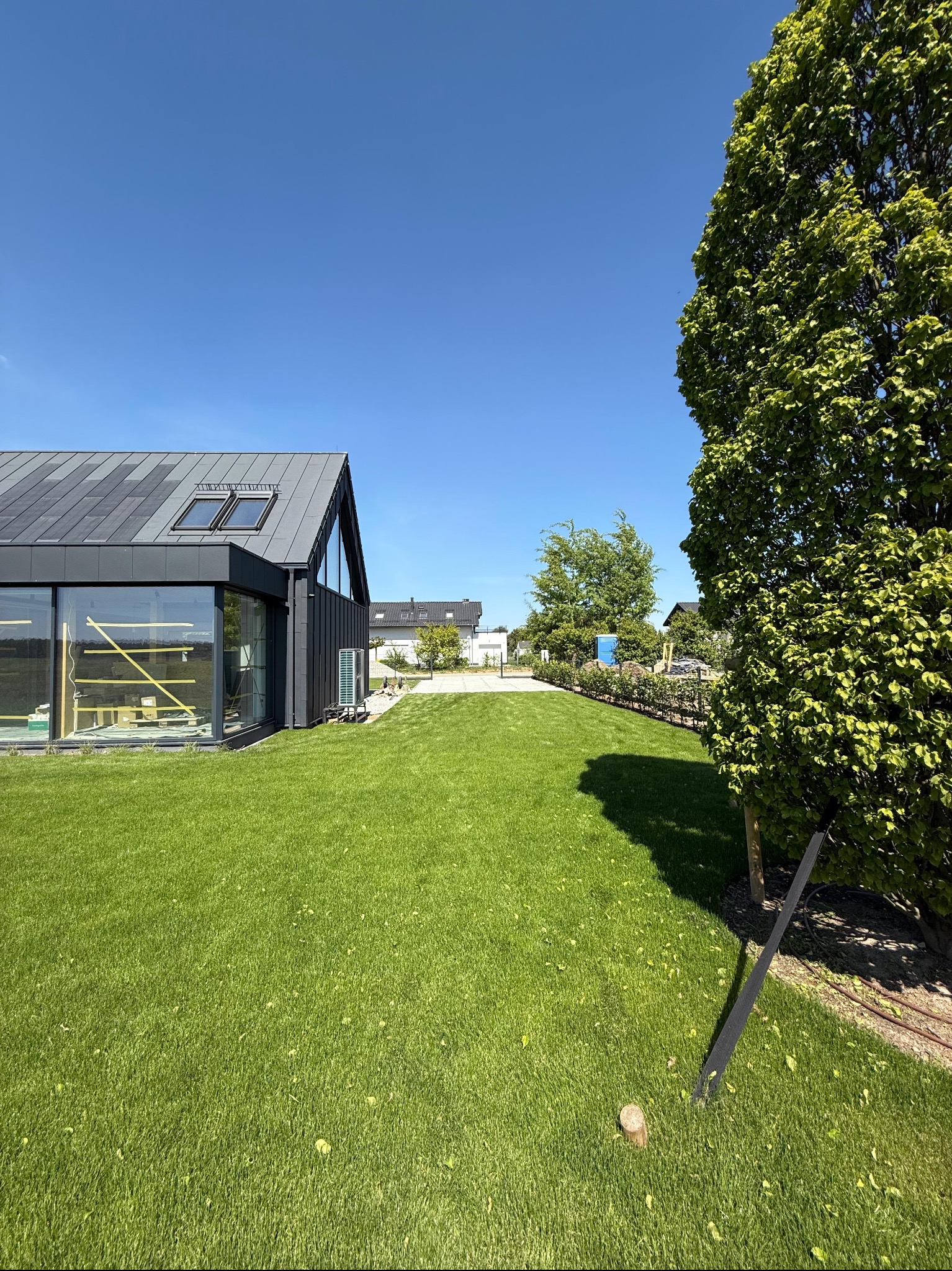 Nowoczesny dom z ciemną elewacją i dużymi oknami, otoczony świeżo założonym trawnikiem i drzewami w słoneczny dzień. Widoczny fragment systemu nawadniania.