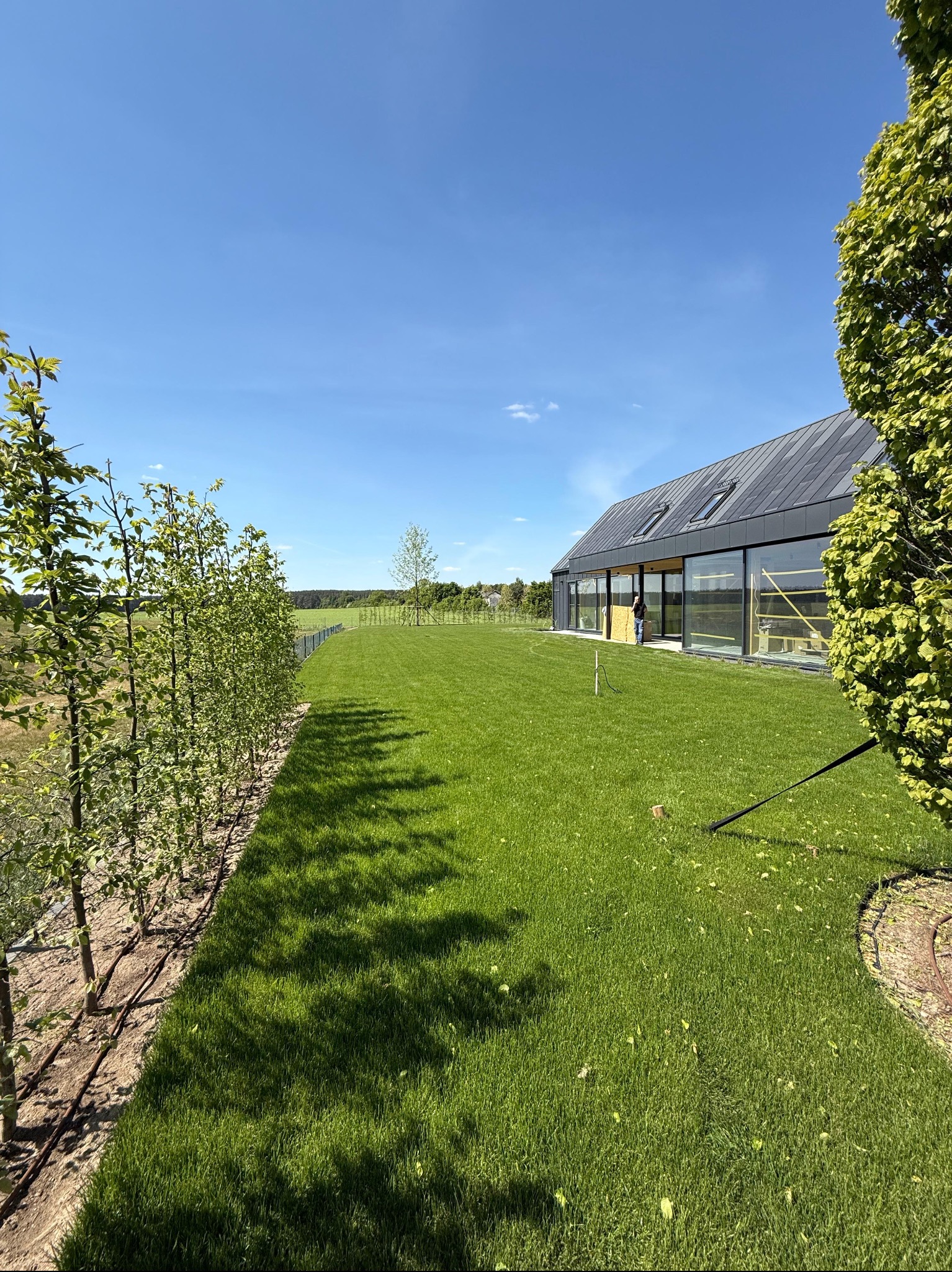 Nowoczesny dom w Gorzowie Wielkopolskim z rozległym trawnikiem, młodymi drzewkami i systemem nawadniania. Błękitne niebo w tle.