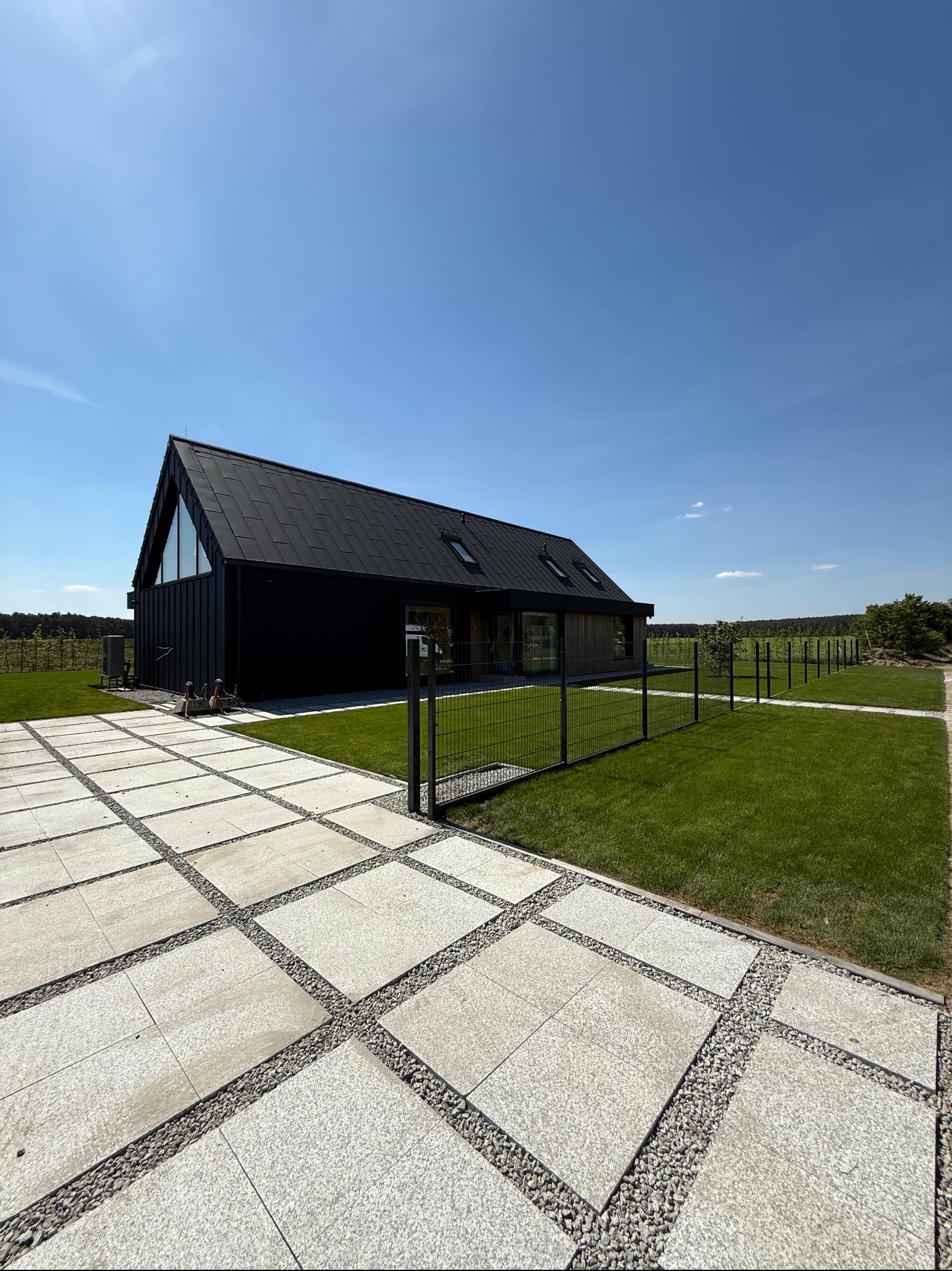 Nowoczesny dom z czarną elewacją, otoczony trawnikiem i ścieżką z płyt. Minimalistyczne ogrodzenie, błękitne niebo. Układ kostki brukowej z drobnym żwirem.