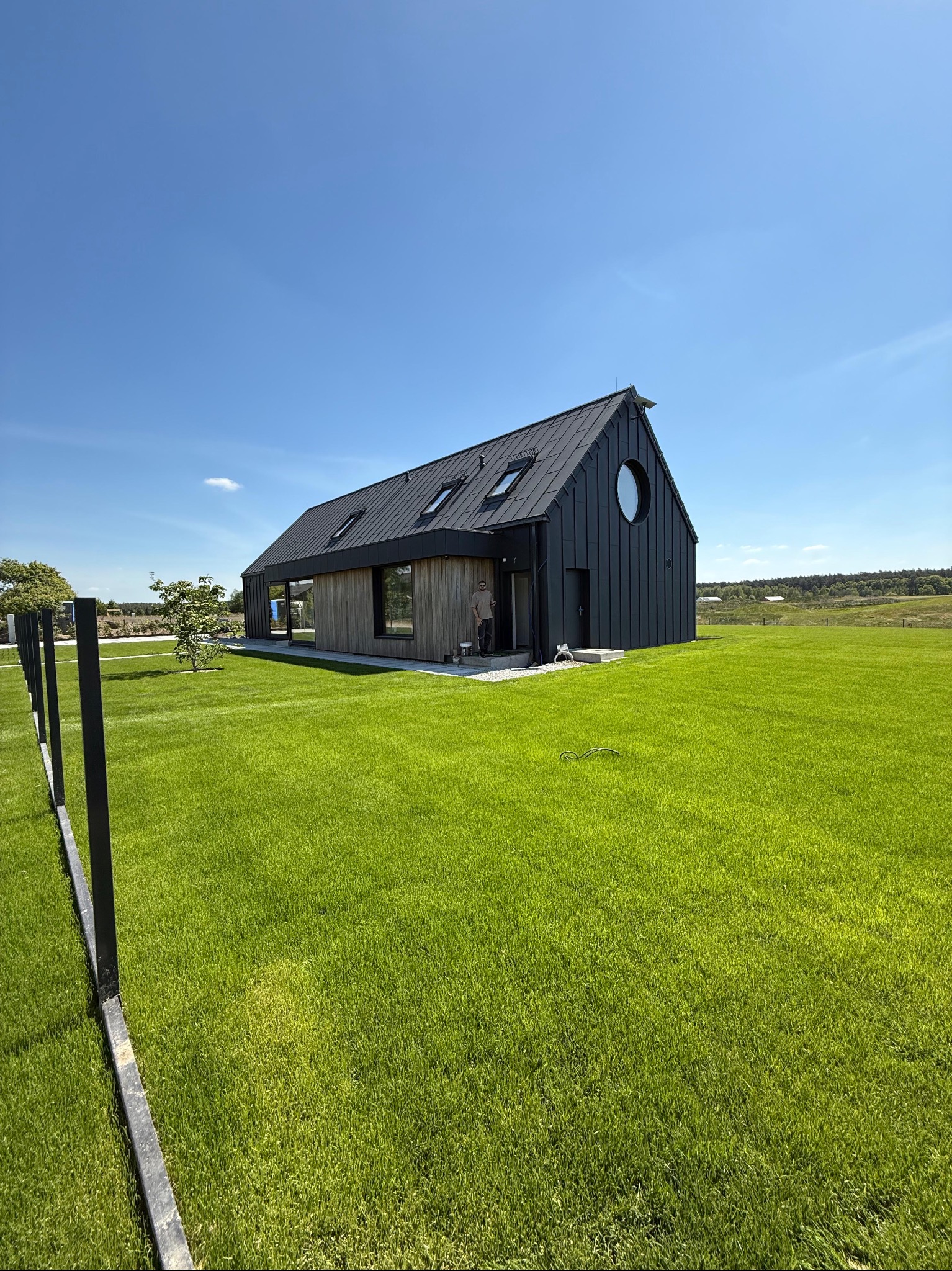 Nowoczesny dom z czarną elewacją i okrągłym oknem, otoczony intensywnie zielonym trawnikiem. W tle błękitne niebo i fragment krajobrazu.